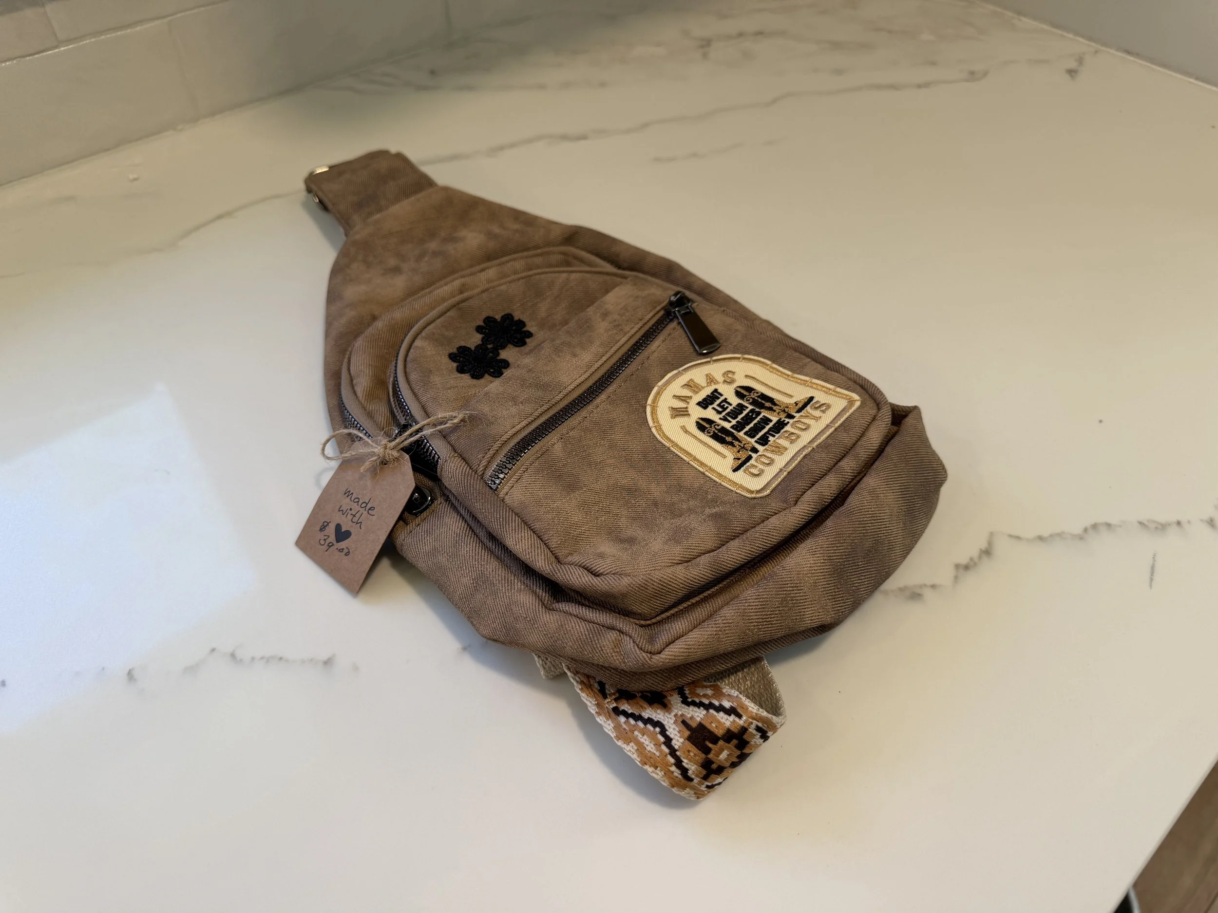 Brown small crossbody bag with decorative patches and a zipper, placed on a white marble countertop.