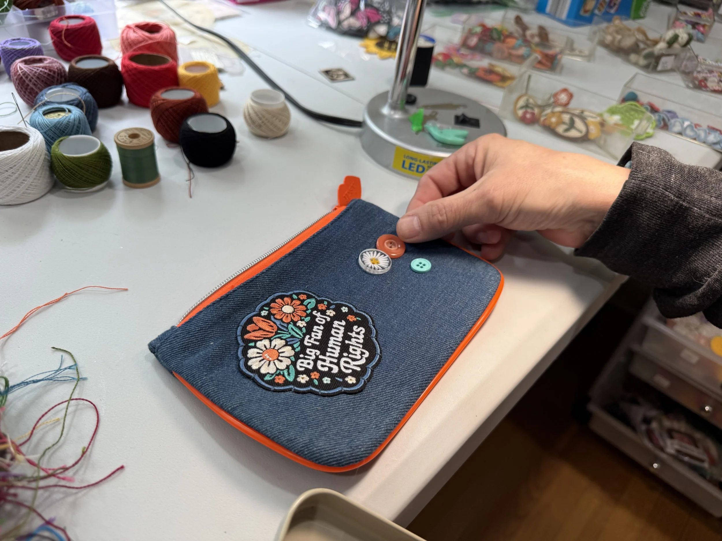 Multiple spools of colorful thread and sewing buttons on a fabric pouch that has embroidered text and floral design on a white table with craft supplies and misc items in the background.