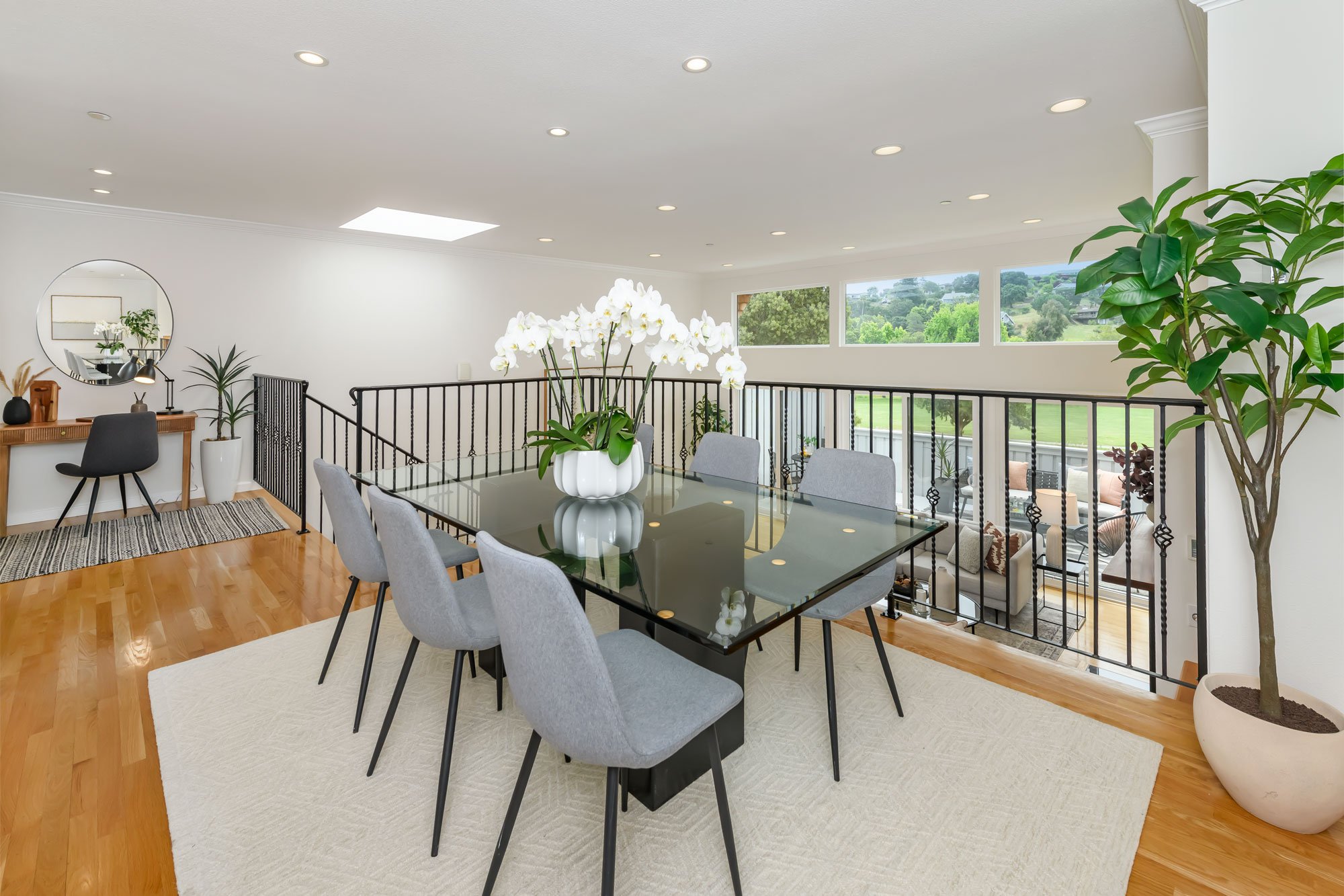 A bright dining area with a glass-top table, surrounded by grey upholstered chairs, center decorated with a white potted orchid. Large windows overlook a green outdoor landscape. The space includes a black railing, a staircase, a wooden console with 