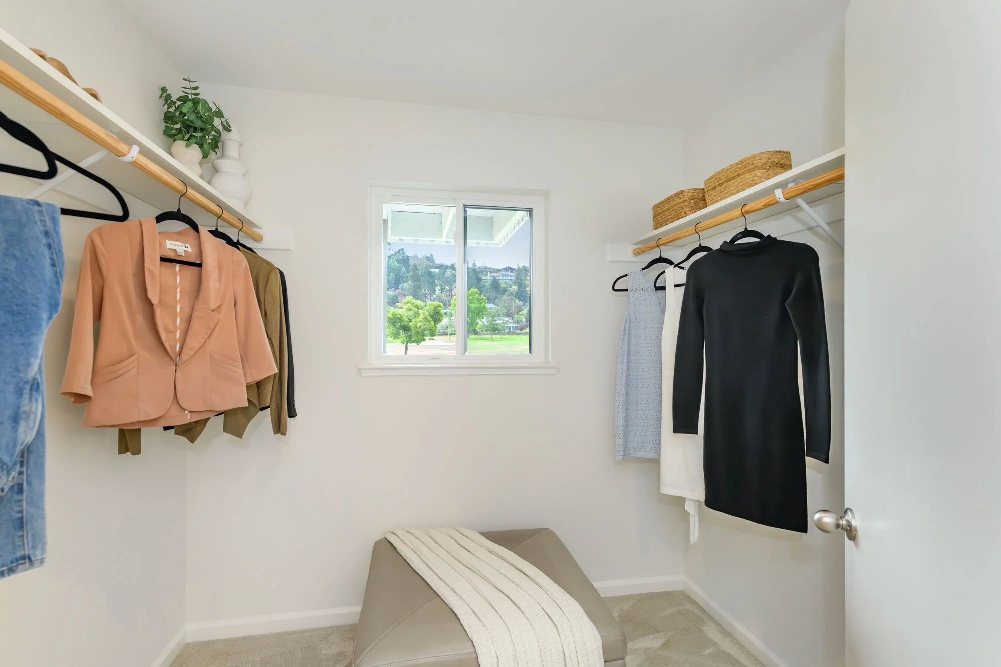 A walk-in closet with white walls, a window looking out onto a green landscape, and a beige cushioned bench. Clothes, including a peach blazer, white dress, black dress, and blue jeans, are hanging on wooden rods, and woven baskets are on the top she