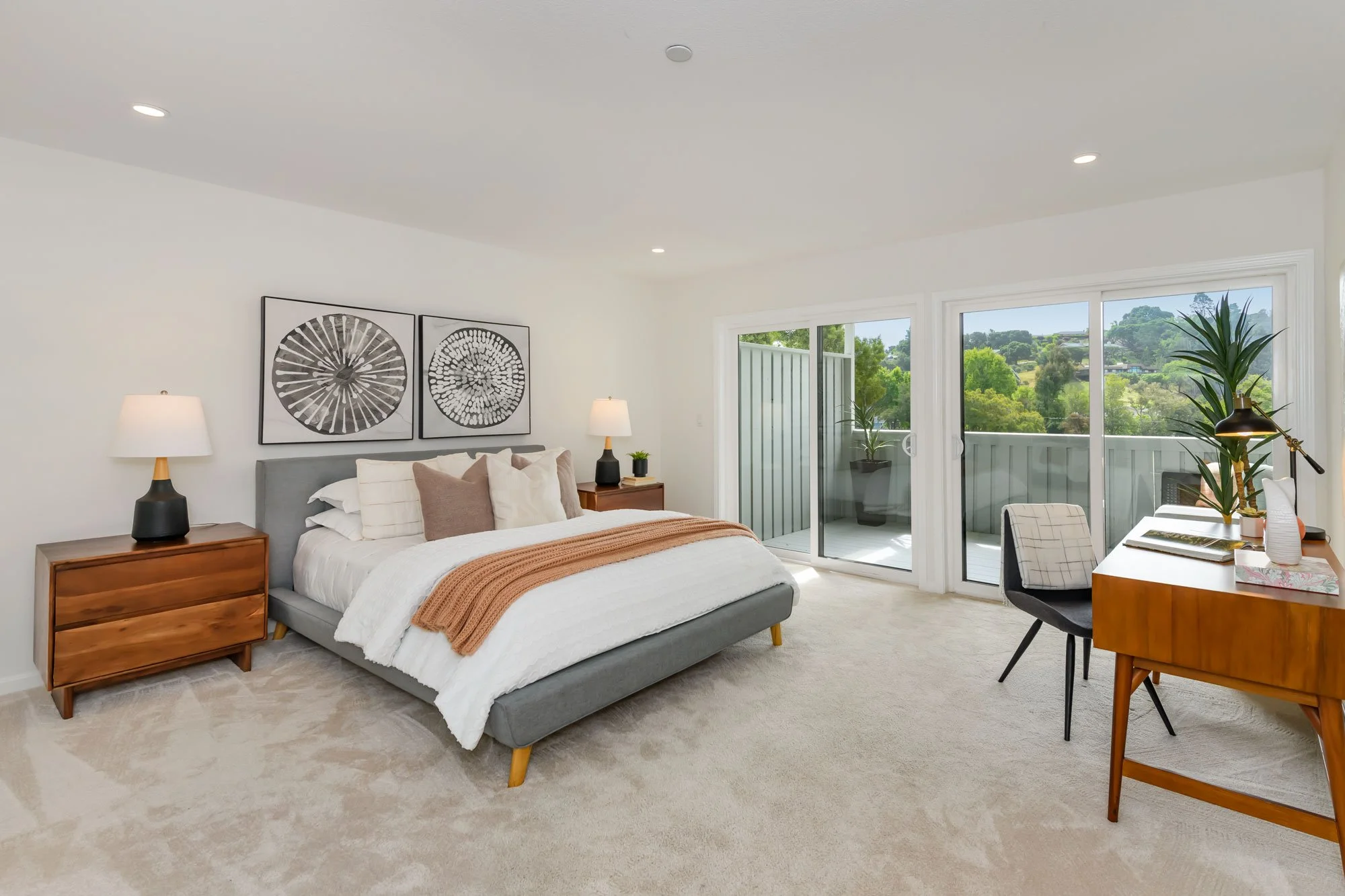 Modern bedroom with large sliding glass door leading to a balcony. The room has a bed with white and beige bedding, wooden nightstands with lamps, wall art, a desk with a chair, and potted plants.