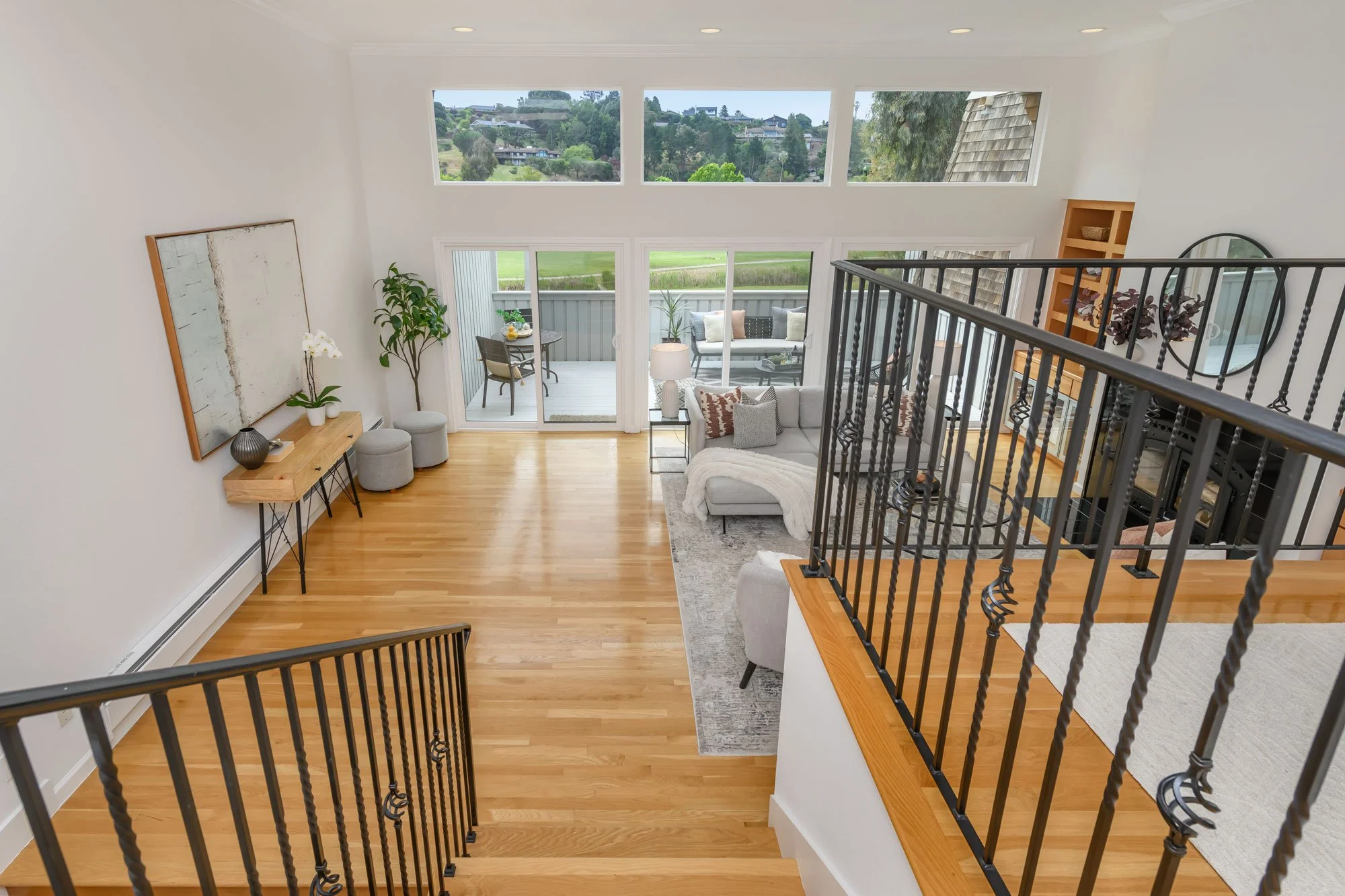 Living room with large windows, sliding glass door leading to outdoor patio, gray sofa with patterned pillows, wooden floor, black metal stair railing, round mirror, and decorative plants.
