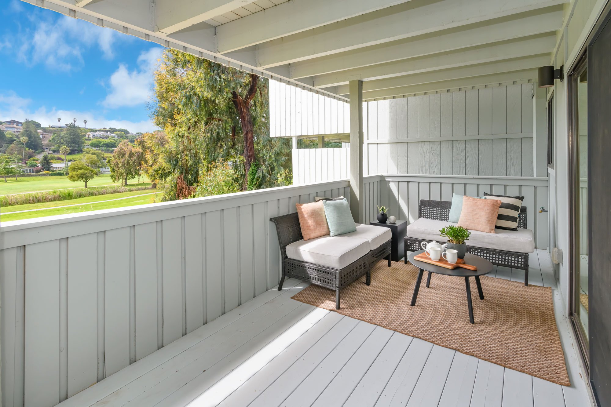 Balcony with white wooden railing and ceiling, outdoor view of lush green park, trees, and distant houses, furnished with black wicker loveseat and chair with throw pillows, small table with tea set, and decorated with potted plant, on a beige rug.