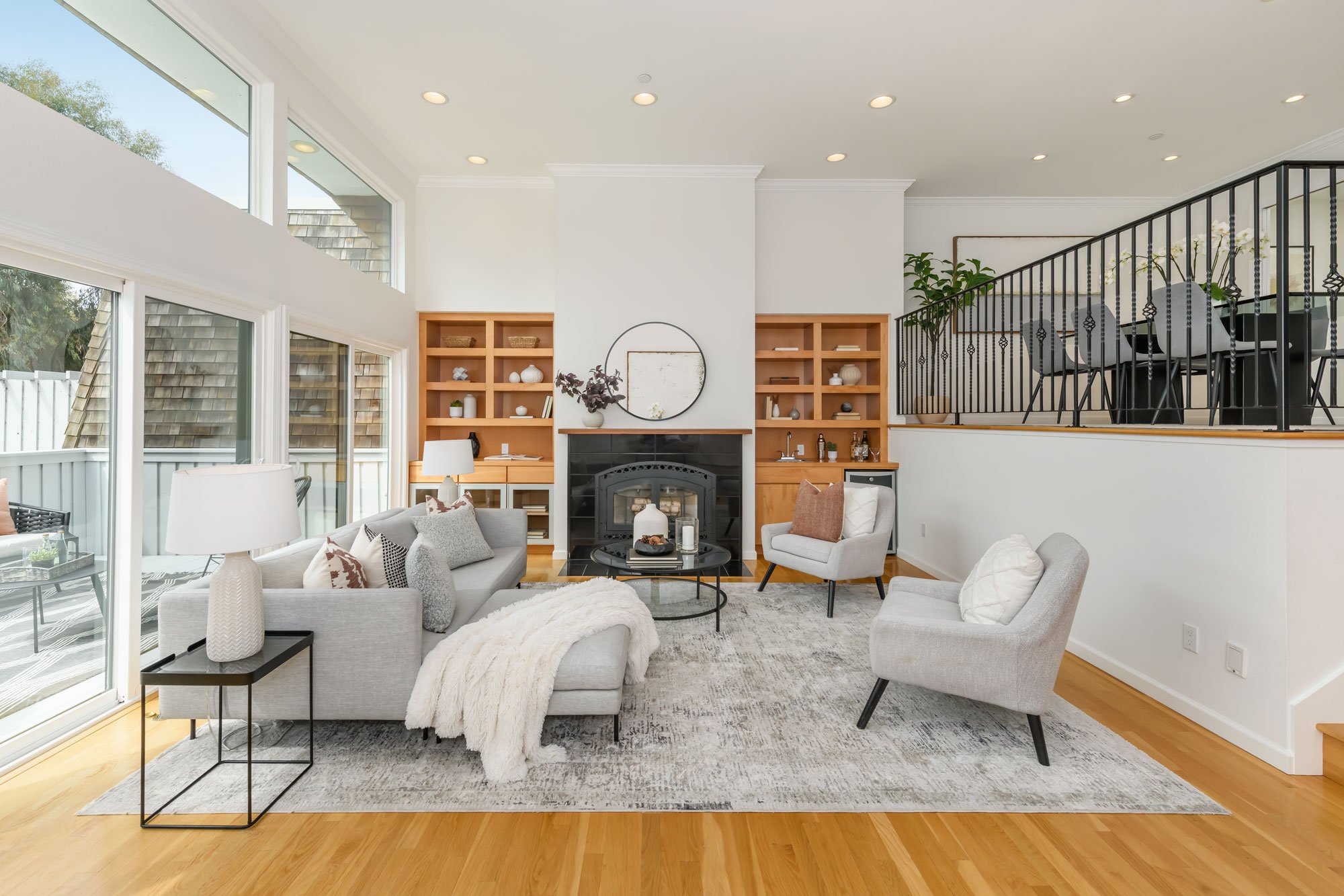 A spacious living room with large windows, white walls, light wood flooring, and a fireplace surrounded by built-in wooden shelves. The room features modern furnishings, including a gray sofa with pillows, a white armchair, and a black round coffee t
