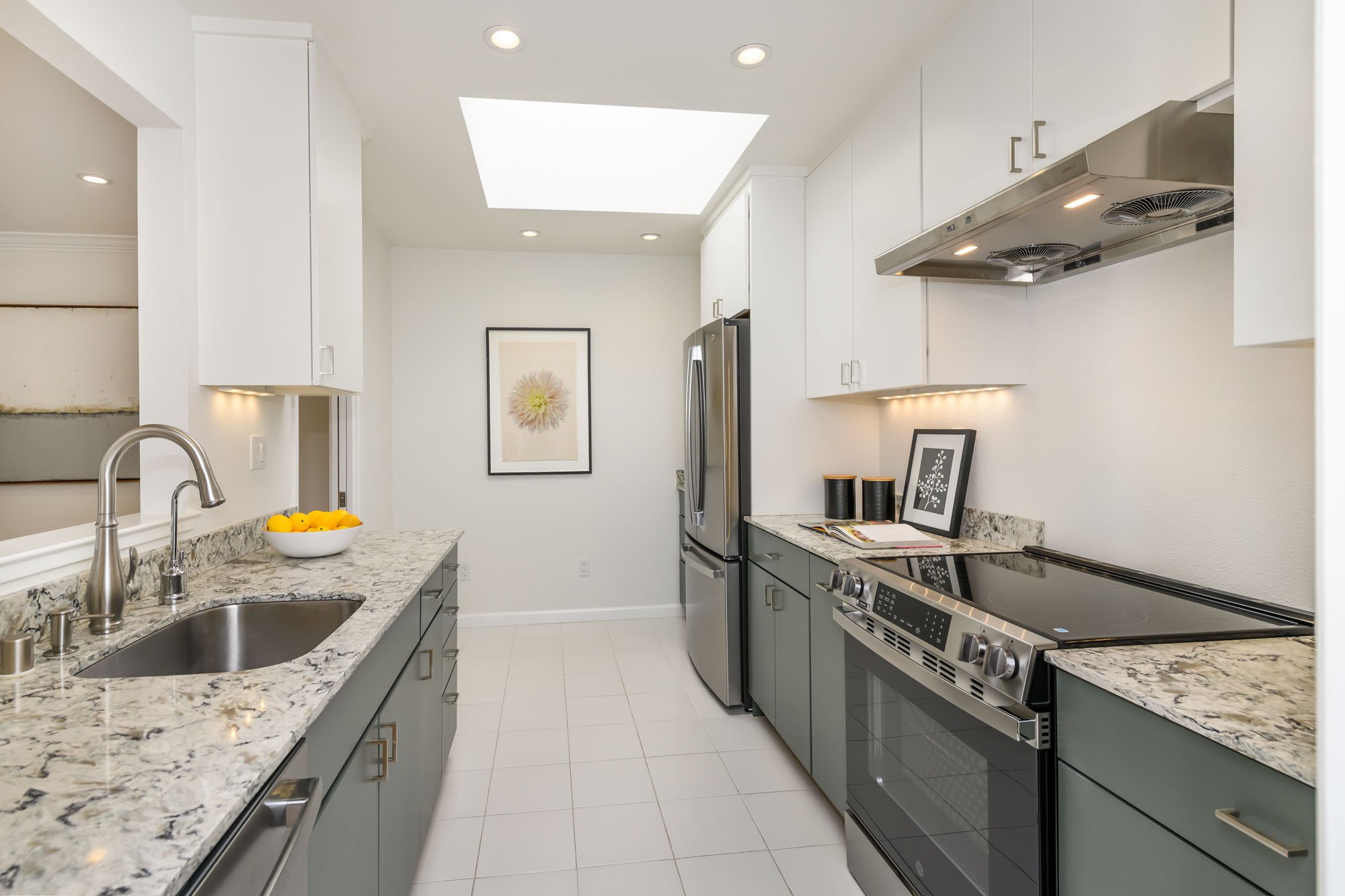 Modern kitchen with white walls and ceiling, skylight, gray lower cabinets, white upper cabinets, stainless steel appliances, marble countertops, and decorative framed artwork, including a bowl of lemons on the counter.