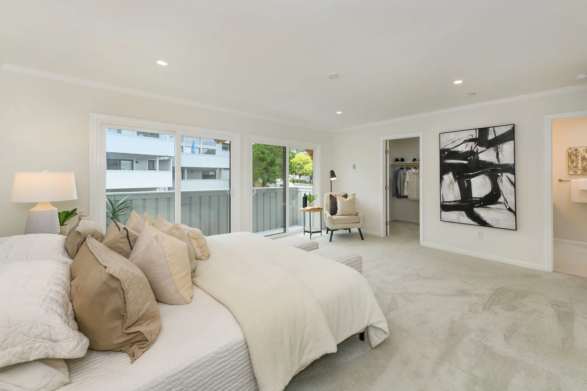 A bright, cozy bedroom with a large bed covered in cream-colored linens and multiple pillows. There is a white table lamp and a beige armchair with cushions near sliding glass doors that lead to a balcony. Outside, apartment buildings and trees are v