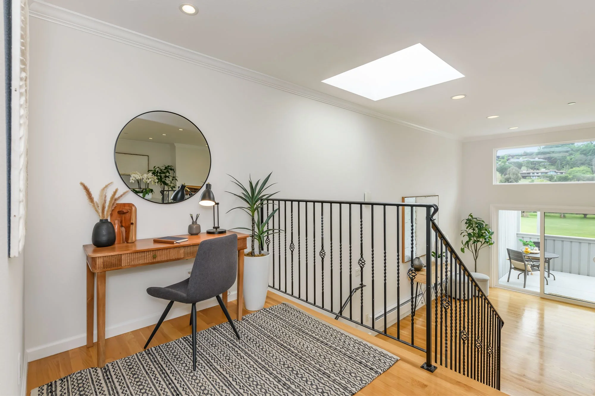 Interior view of a modern home with a small desk, round mirror, potted plants, a desk lamp, a chair, wooden flooring, and a staircase railing leading to a room with large windows and outdoor view.