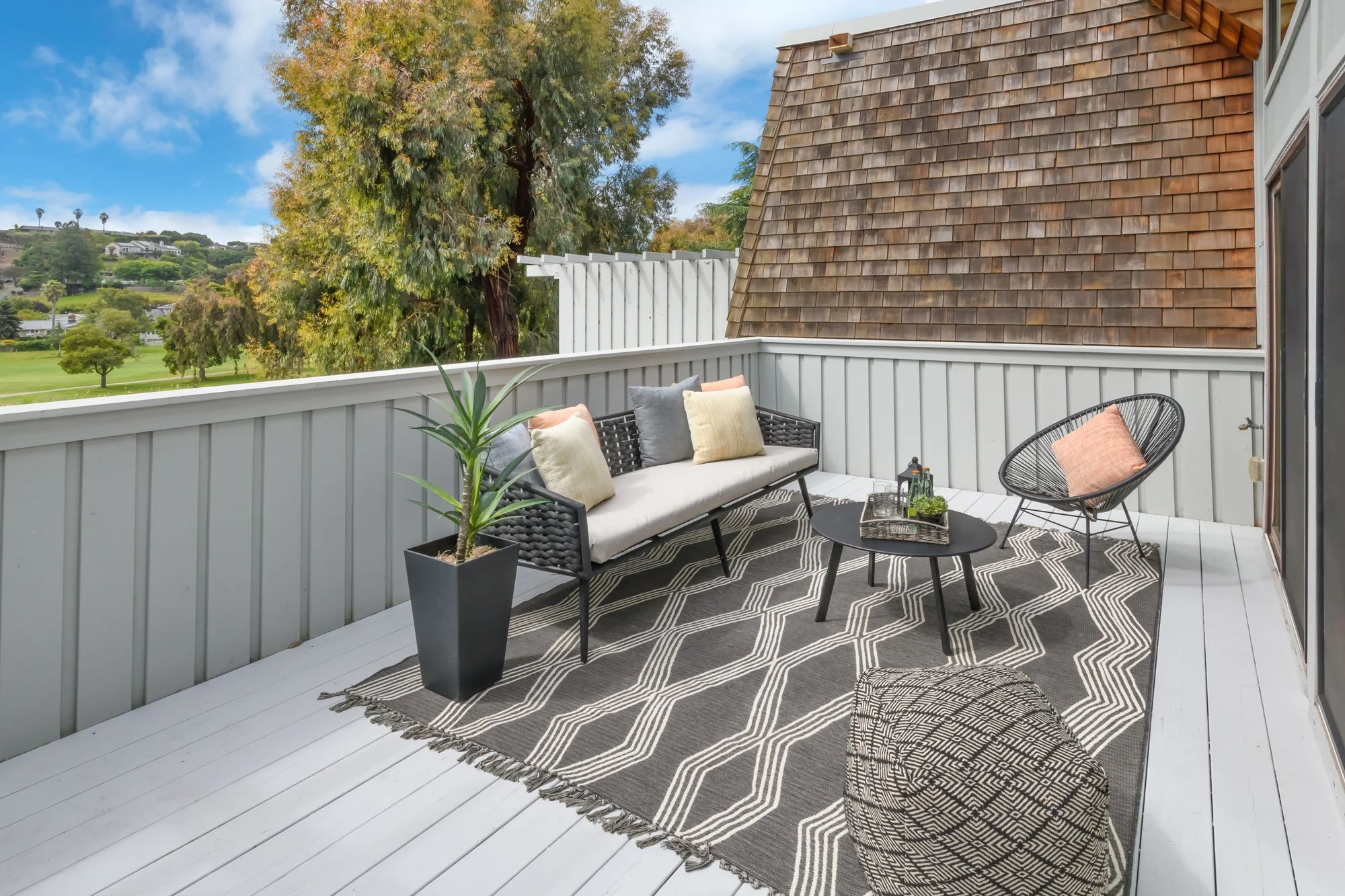 Balcony patio with black wicker sofa with beige, gray, and pink cushions, black round coffee table with decorative items, black and white patterned rug, gray metal planter with green plant, black woven chair with peach cushion, view of green park, tr