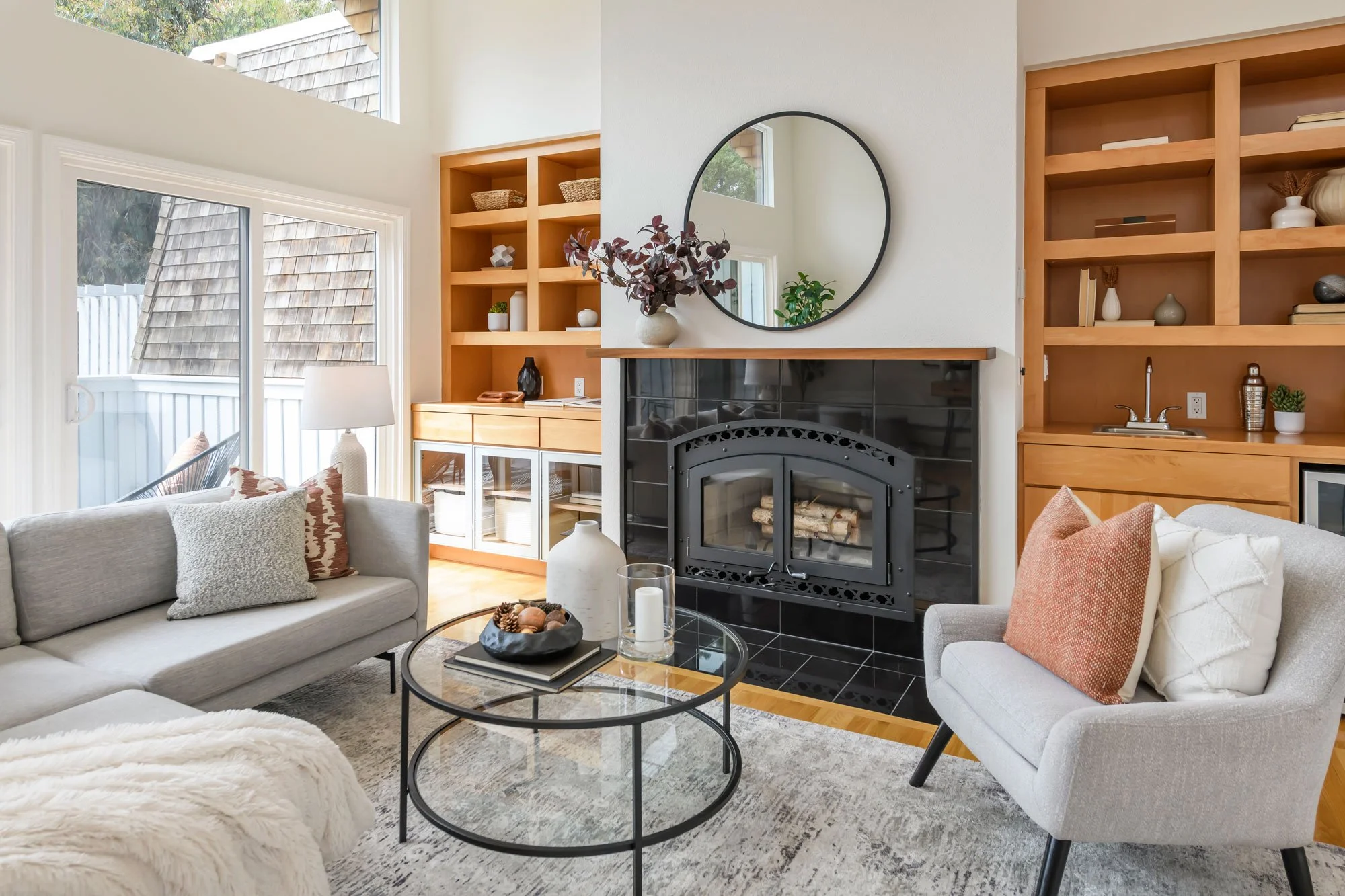 Modern living room with light-colored sofas, a glass coffee table with decorative items, a black fireplace, and wooden shelves with decorative objects. Large windows let in natural light.