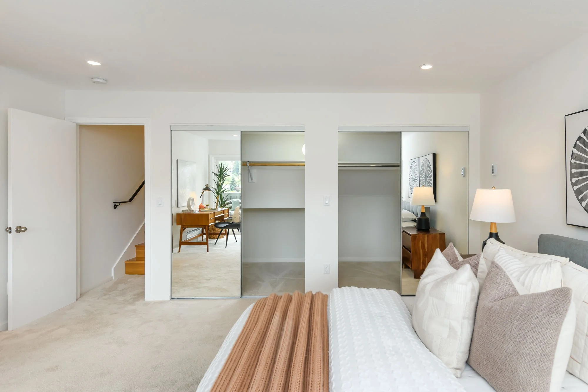 A bedroom with a bed, pillows, and a blanket, with two bedside lamps, and a large sliding mirror closet door. The room opens to a hallway and another room with a desk, chair, and window with a plant.