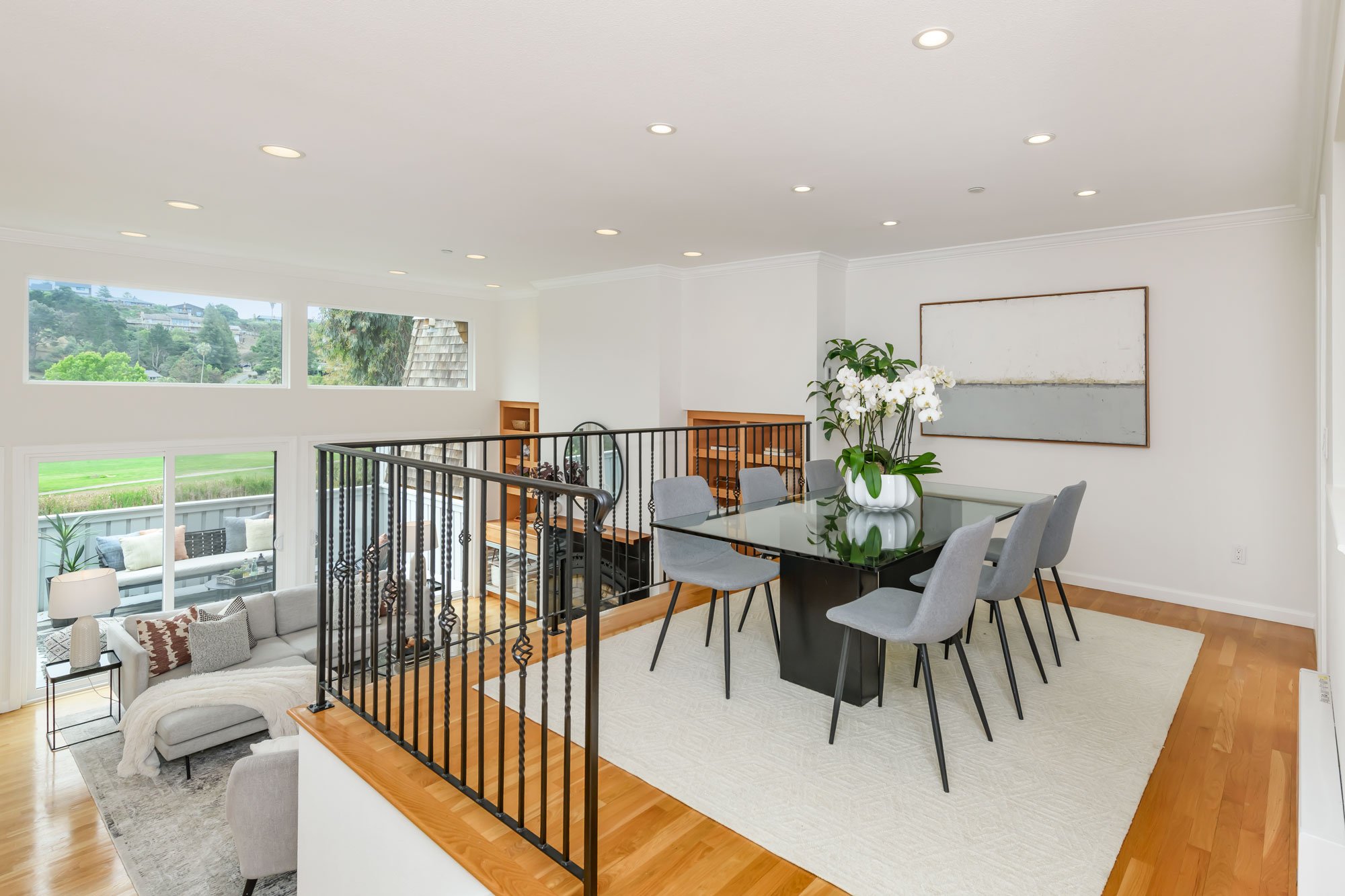 A spacious, well-lit modern open-concept living and dining area with large windows, white walls, and light hardwood floors. There is a dining table with light gray chairs, a glass door, a room with a white sofa and decorative pillows, and a large abs