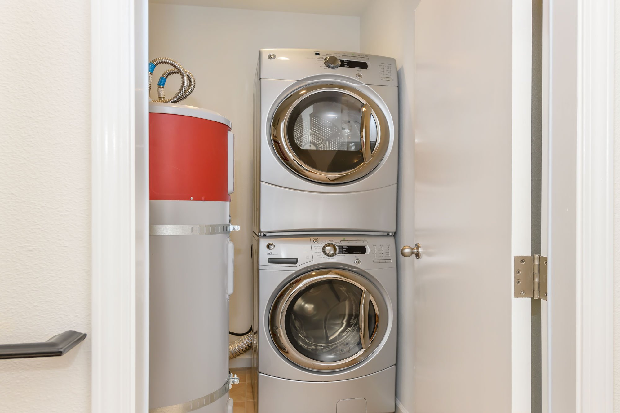 A small laundry room with a stacked washer and dryer, a red water heater to the left, and beige walls.
