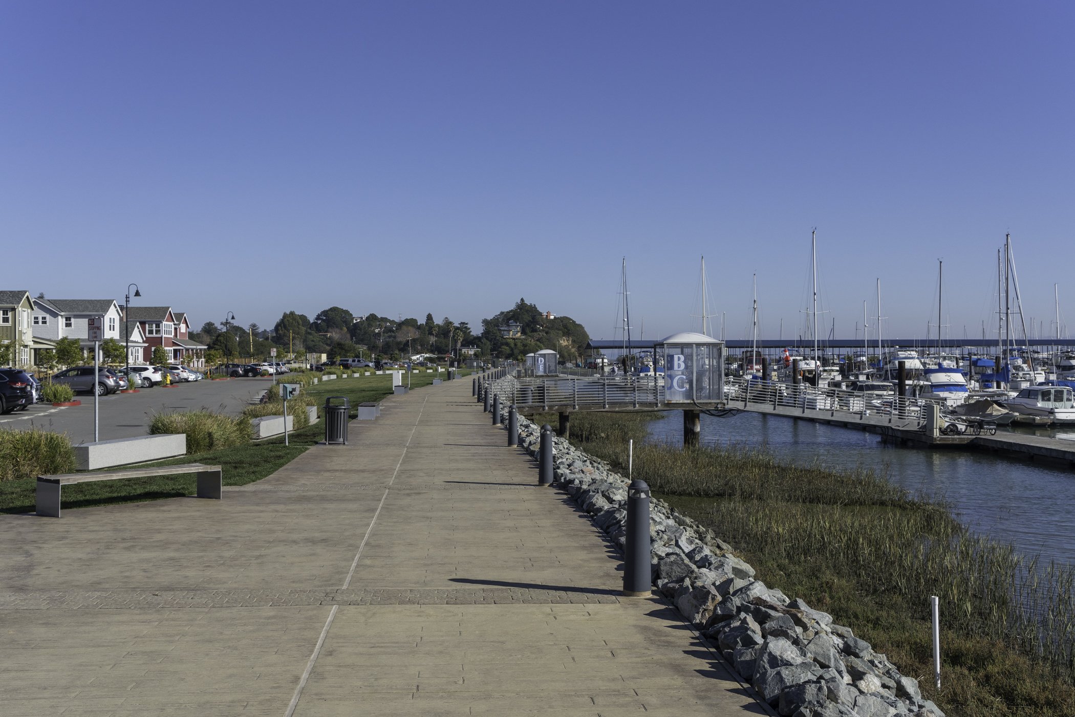 A paved walkway by a marina with boats docked on the right and colorful houses on the left under a clear blue sky.