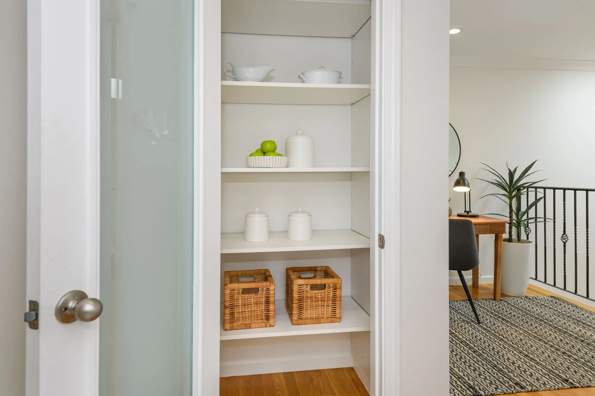 Empty white open shelving closet with baskets, ceramic jars, and bowls, viewed from a doorway. In the background, part of a living area with a black plant in a white pot, a wooden side table, and a black and white patterned rug.