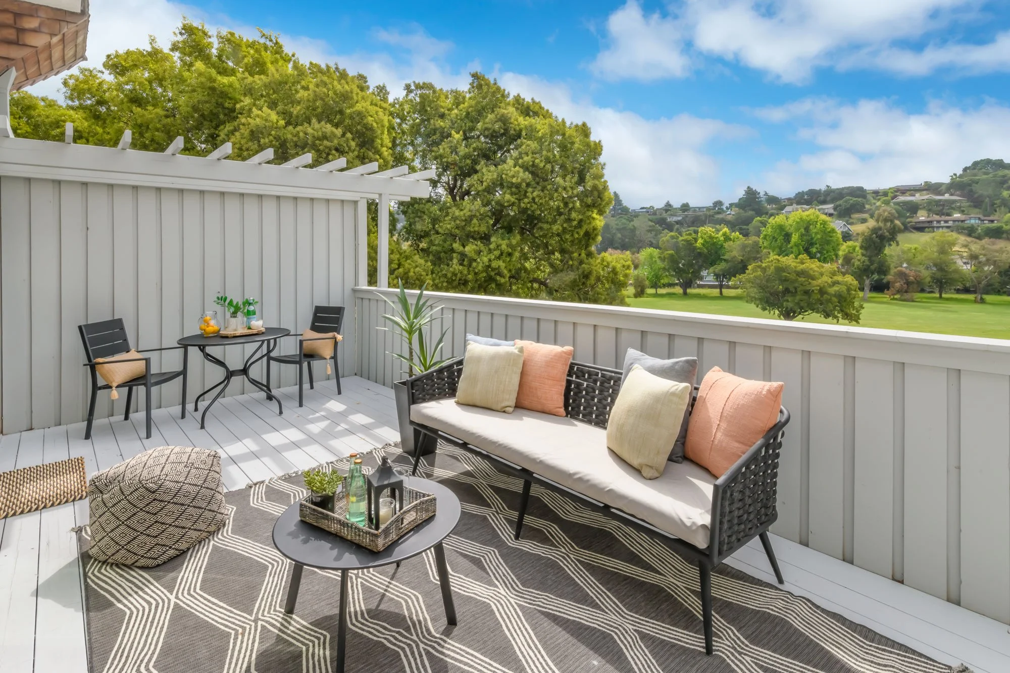 A balcony with a white wooden railing, black patio furniture including a loveseat, chairs with cushions, a small round table with a tray of drinks and plants, a patterned outdoor rug, and a large green tree with hills and houses in the background und