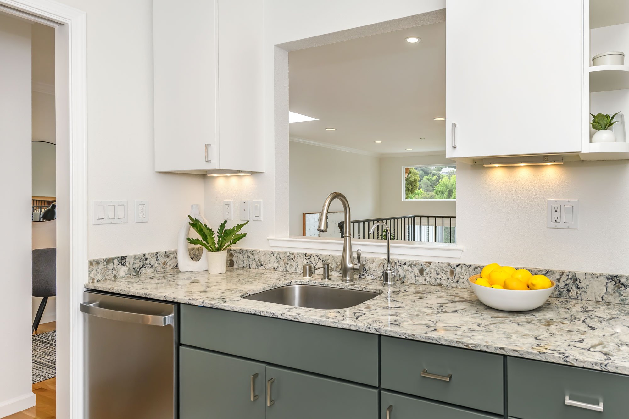 Kitchen with granite countertop, white upper cabinets, gray lower cabinets, stainless steel sink, faucet, a bowl of lemons, and a potted plant. Open view of the living room and window showing greenery outside.