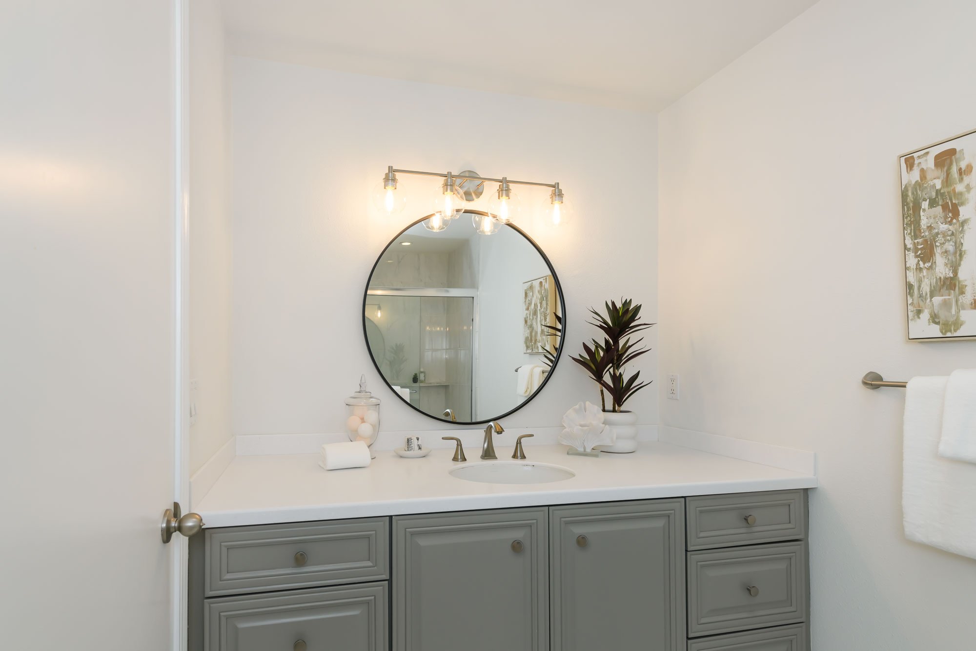 A modern bathroom vanity with a round mirror, light fixture above, and decorative plants and accessories on countertop, white walls, and a piece of artwork on the wall.