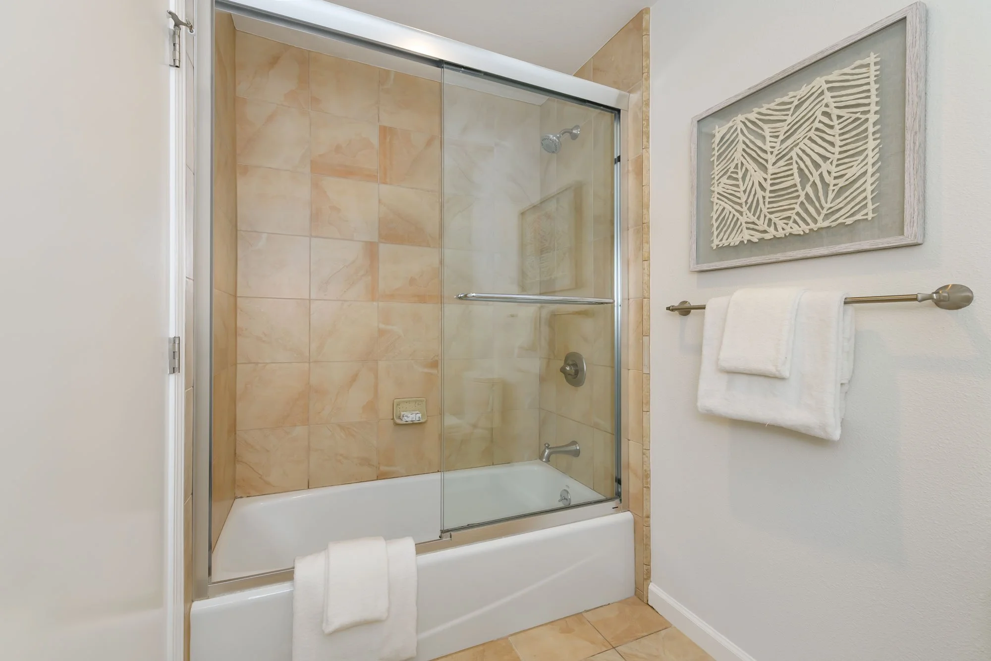 Bathroom shower with glass door, beige tiled walls, a white bathtub, white towels on a silver towel rack, and a framed piece of artwork on the wall.