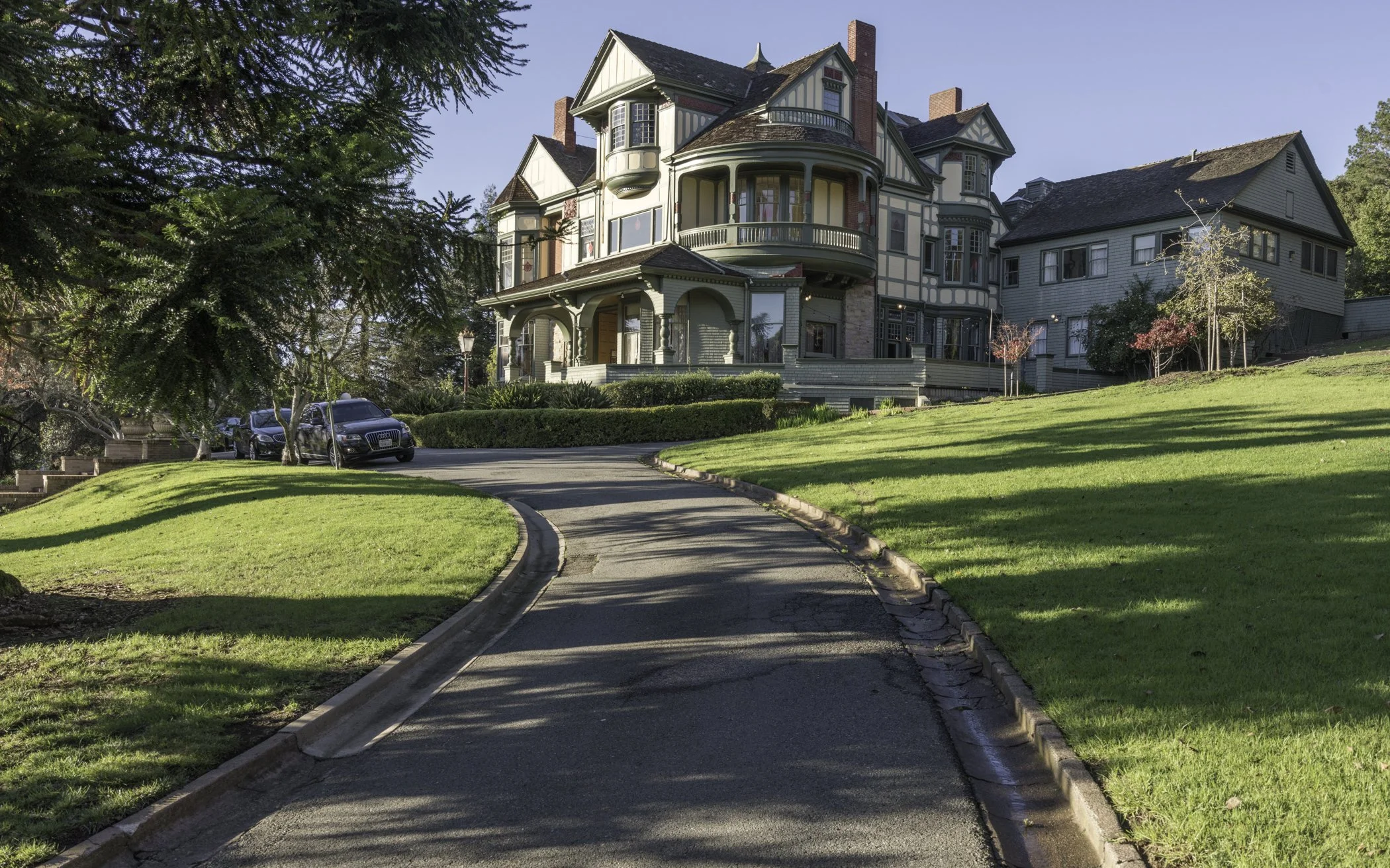 A large, Victorian-style house with multiple stories, bay windows, and a wrap-around porch, surrounded by green lawns and trees. A curved driveway leads up to the house, with parked cars visible on the left side. The sky is clear and blue.