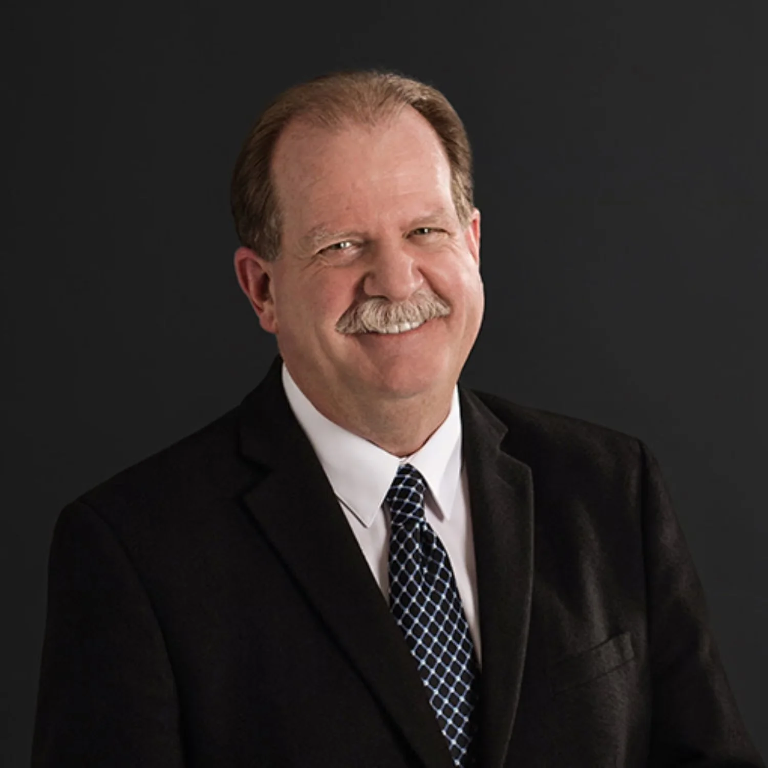 Portrait of a smiling man with a mustache, wearing a black suit, white shirt, and patterned tie, against a dark background.