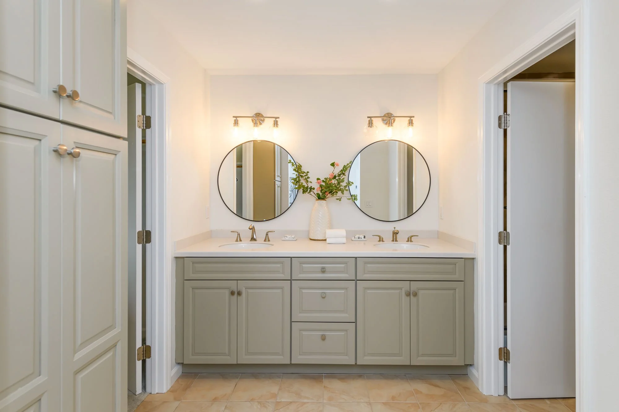 A bathroom vanity with dual sinks, beige cabinets, two oval mirrors, and wall-mounted lights. A vase with pink flowers and a rolled towel are on the countertop. Two open doorways are on either side.