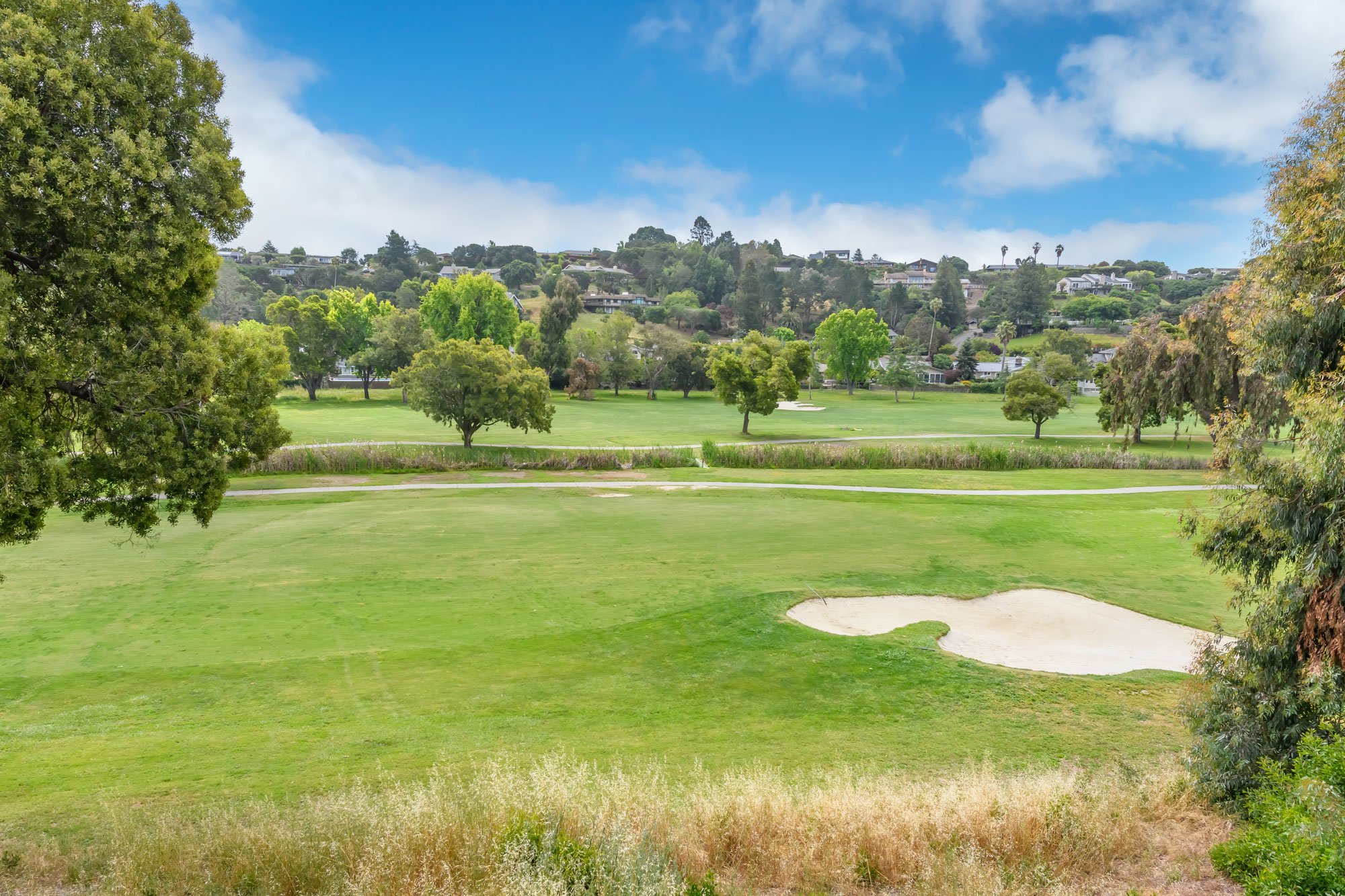 A golf course with green grass, sand bunkers, trees on the course and in the surrounding areas, houses on a hillside in the background, blue sky with clouds.