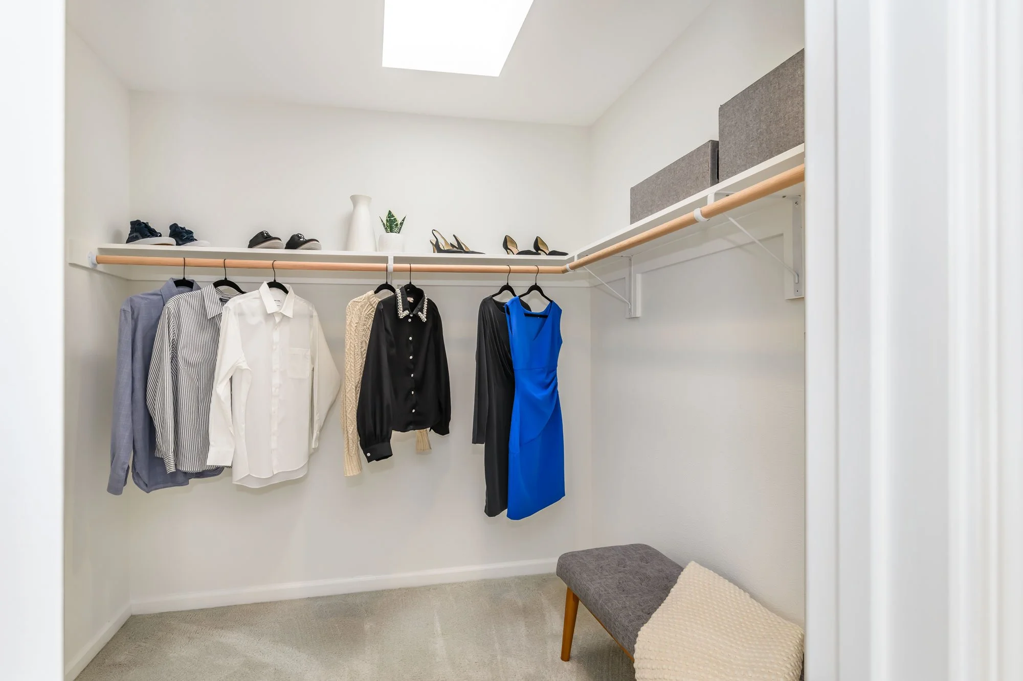 A walk-in closet with a hanging rod holding various clothing items including a gray shirt, a white shirt, a cream sweater, a black shirt, and a blue dress. The top shelf displays shoes, a white vase, a potted plant, and three storage boxes. is a smal