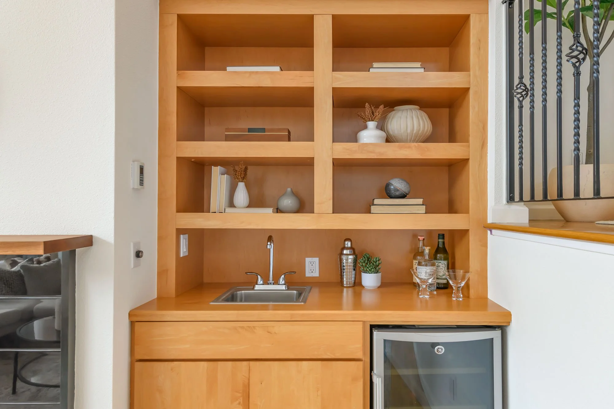 Wooden kitchen shelf with decorative items, bottles, plants, a small sink, and a mini refrigerator, next to a staircase.