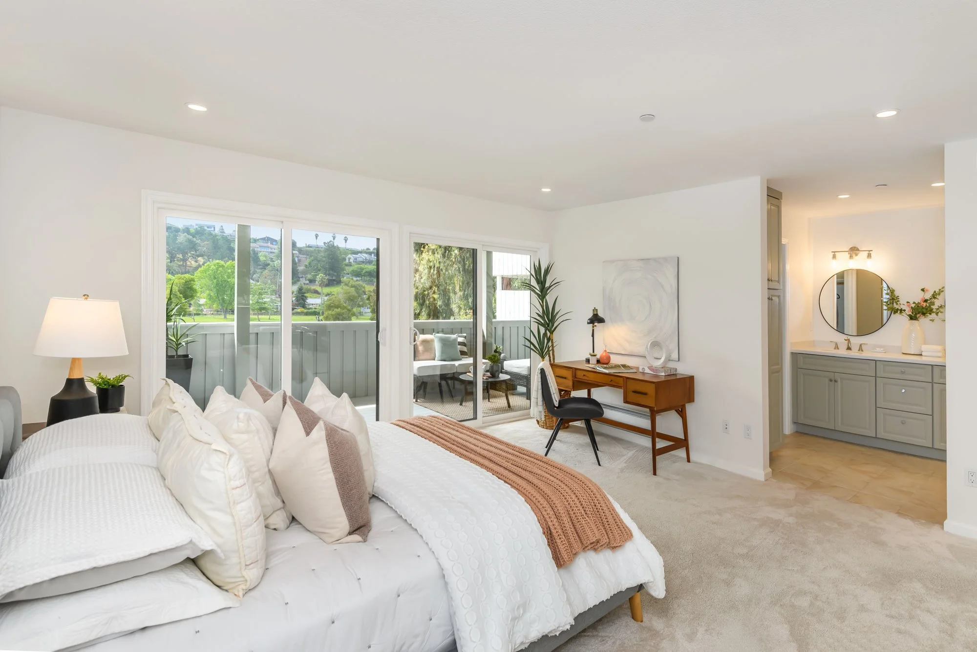Bright, airy bedroom with large windows showing outdoor green scenery, white bedding with multiple pillows, a bedside lamp, a small desk with a chair, and a bathroom with a round mirror and a vase of flowers.