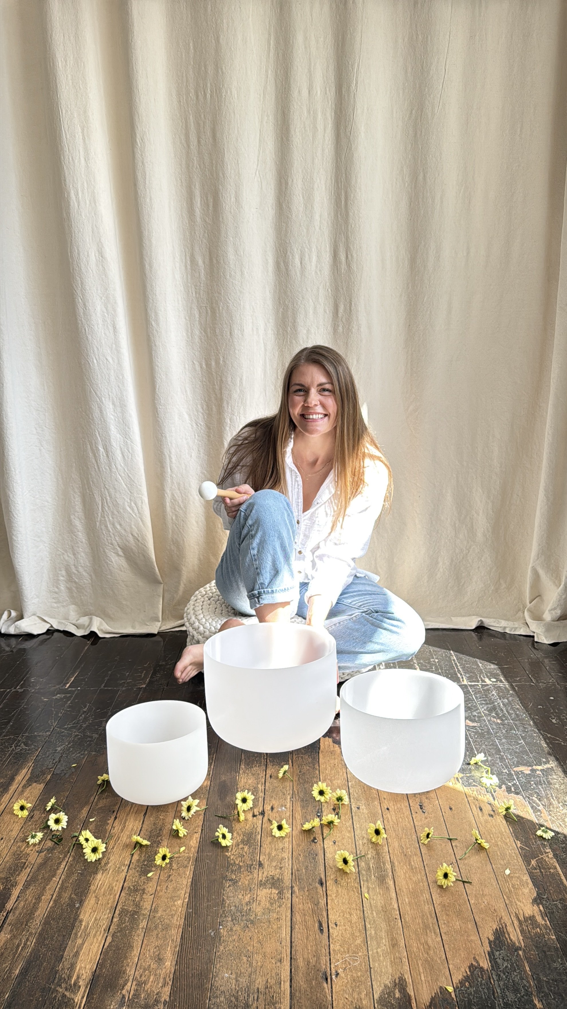 A woman sitting on the floor playing crystal singing bowls, surrounded by yellow flowers, with a cream-colored curtain as a backdrop.