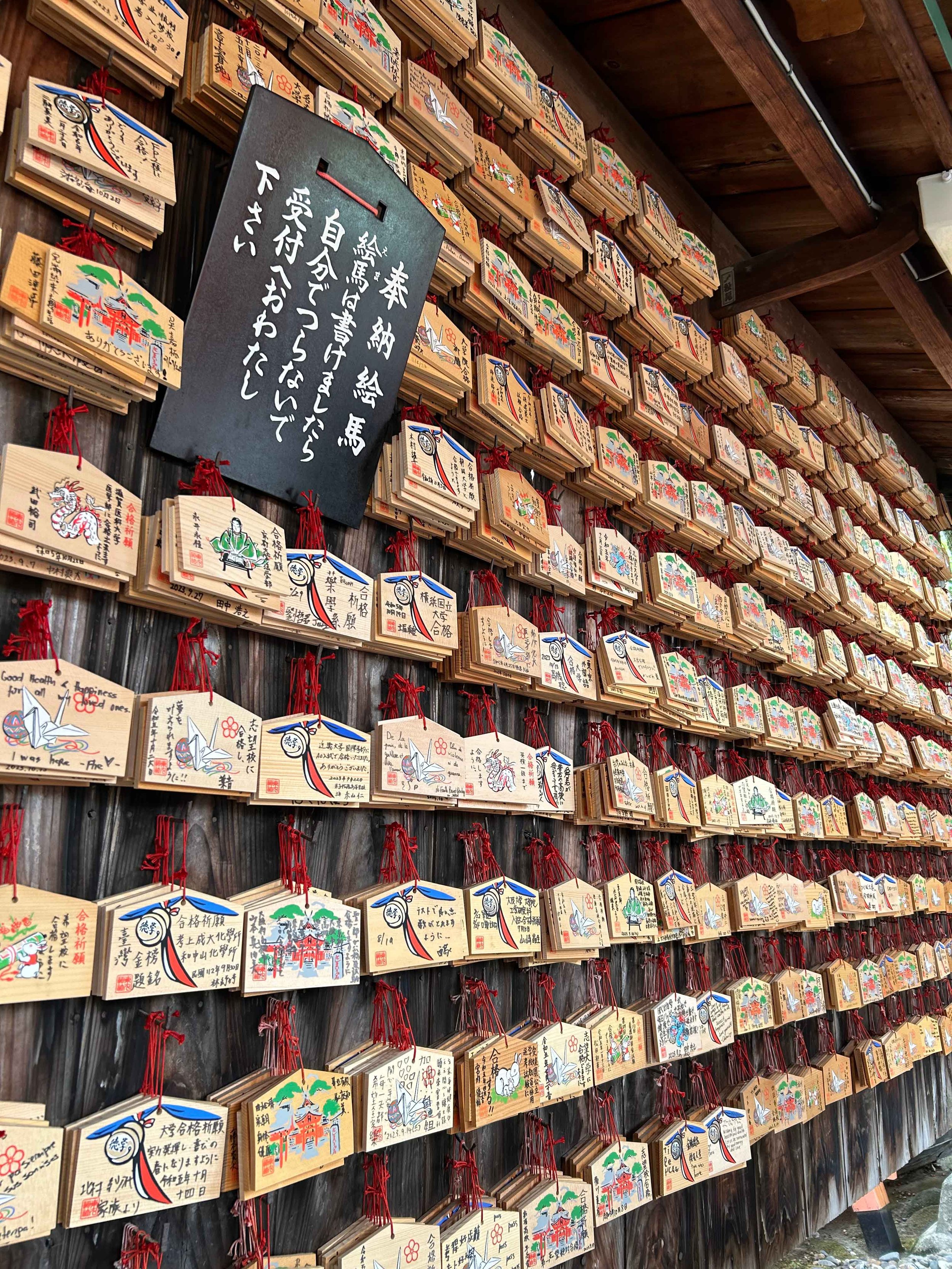 A wall lined with numerous wooden prayer plaques called Ema, each decorated with colorful illustrations and handwritten messages, at a Japanese shrine. Photo taken on the trip to japan in 2023.