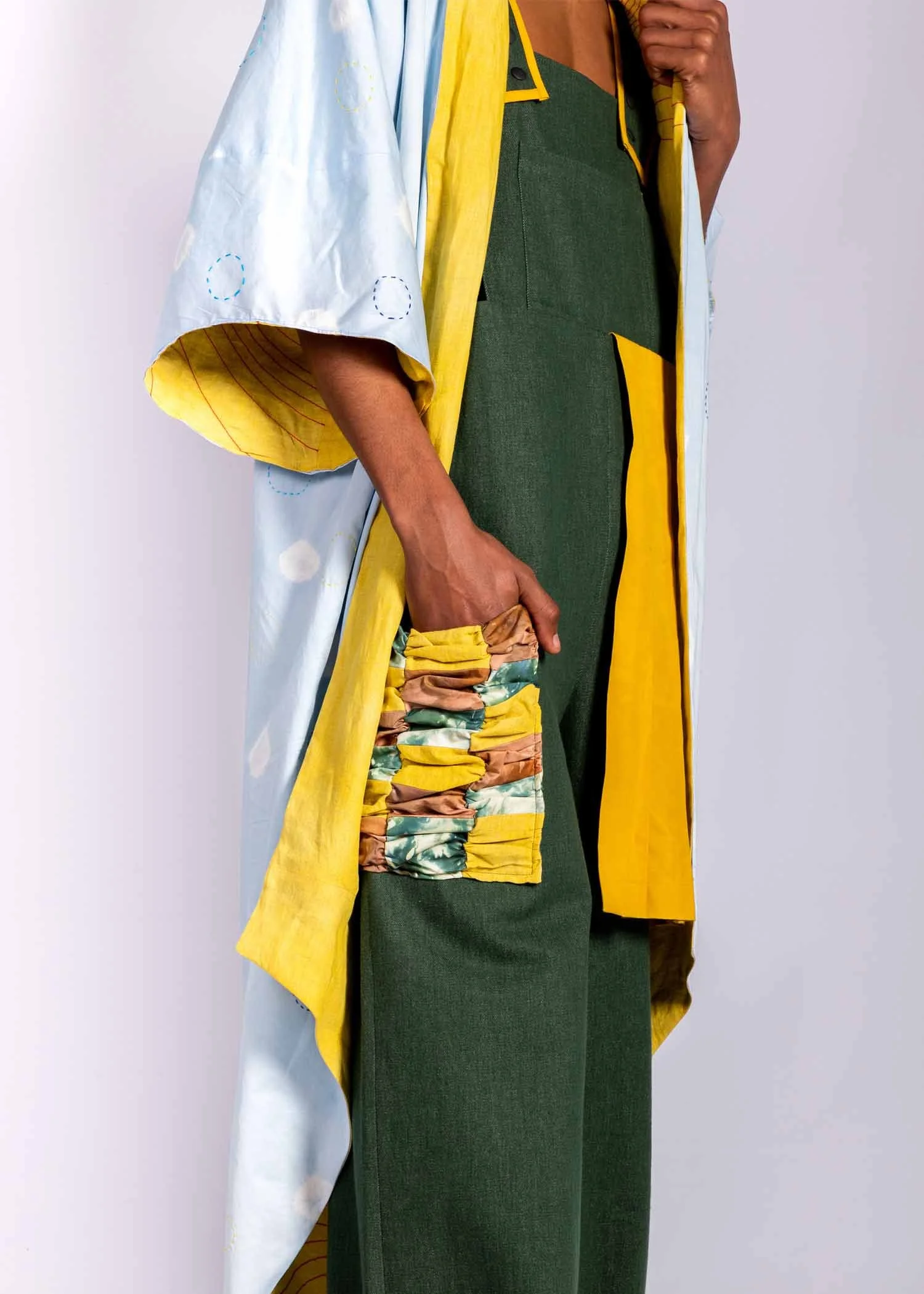 Close-up of a person wearing a multicolored jacket over a green dress, with hand resting on the side and a colorful fabric detail on the dress.
