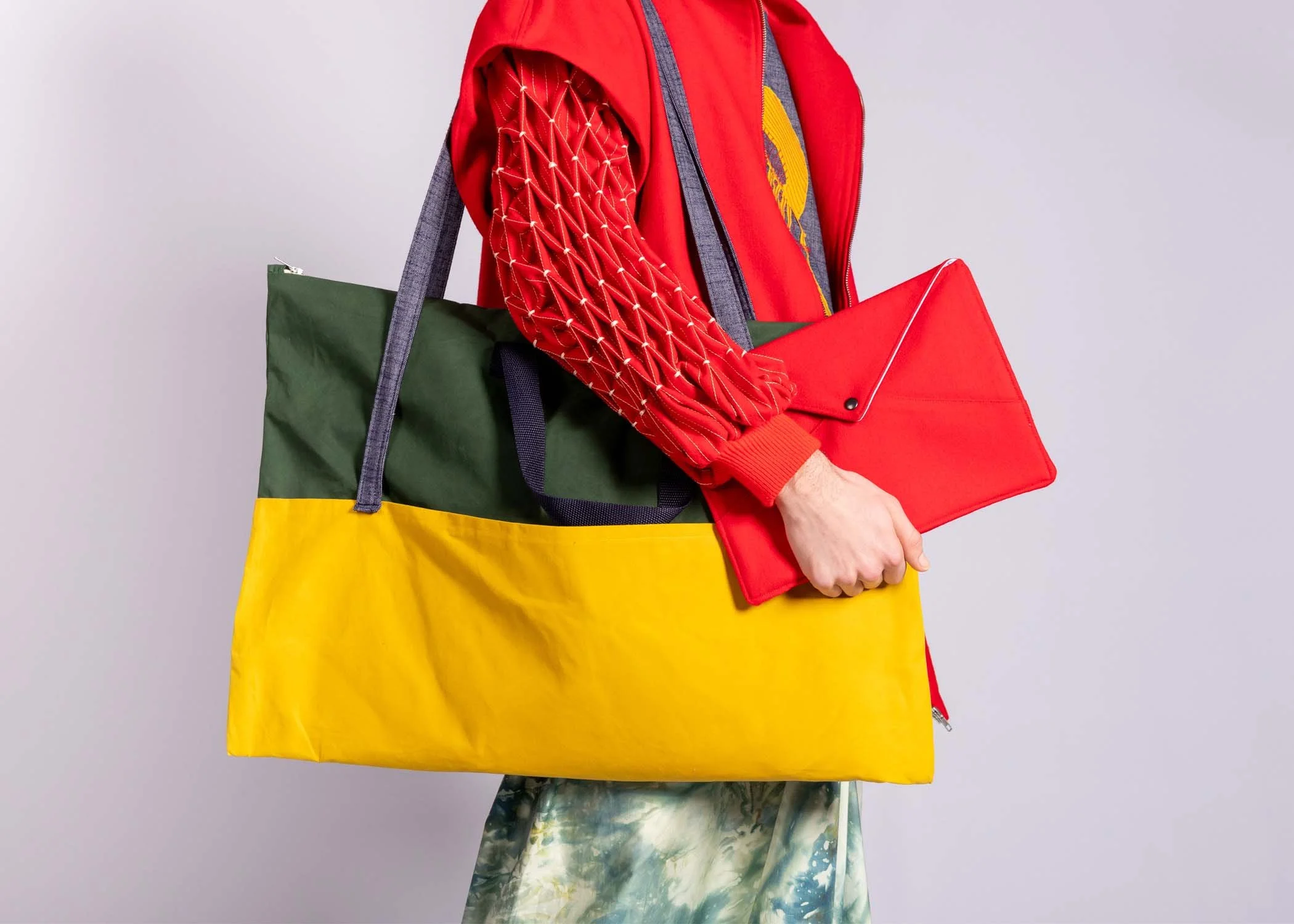 Person wearing colorful clothing holding tote bags and a laptop with a plain gray background.