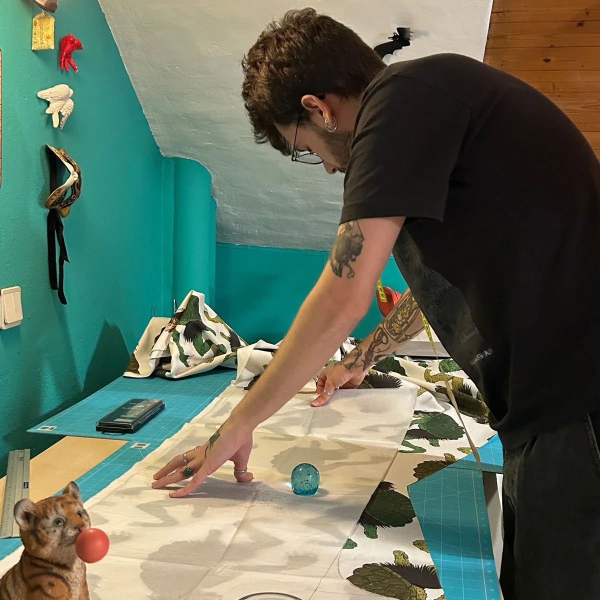 Akai Ito fashion designer, is bending over a table with fabric, a small tiger figurine and glass marble on it, in a room with teal walls and wall decorations, involving sewing or craftwork.