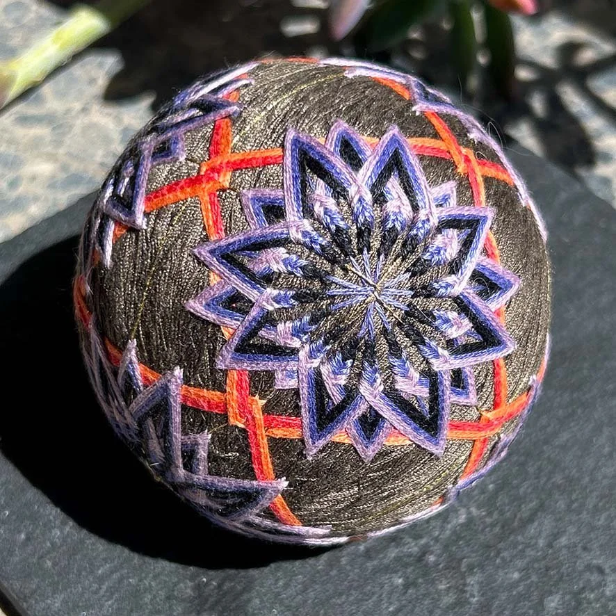 Close-up of a temari ball , fabric waste ball, decorated with intricate embroidered geometric patterns in purple and red thread.