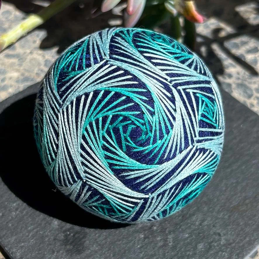 Colorful spherical temari ball with intricate, swirling line patterns in shades of blue, turquoise, and white.
