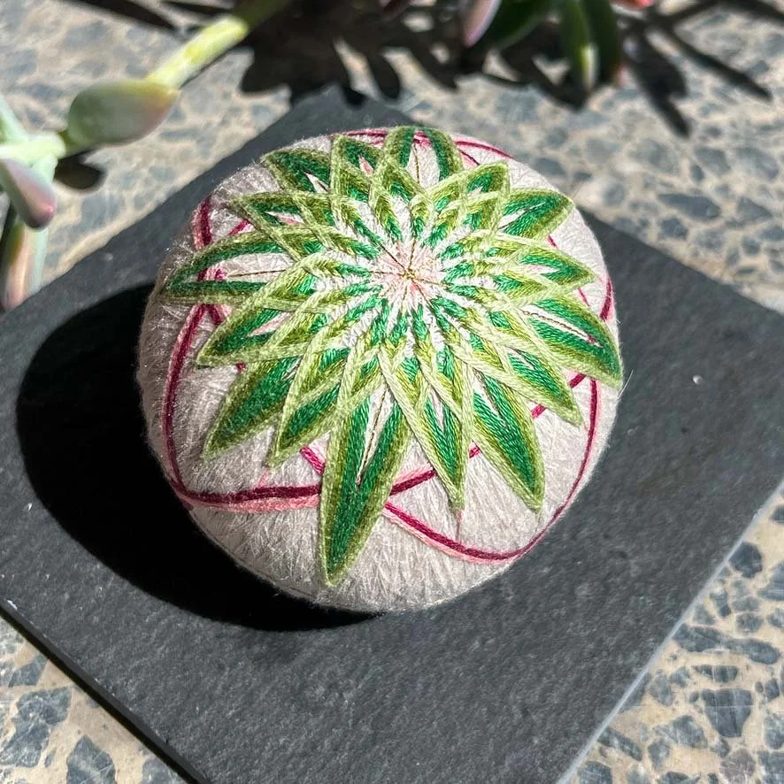 Decorative embroidered green starburst pattern and pink outline temari ball, placed on a black surface outdoors.