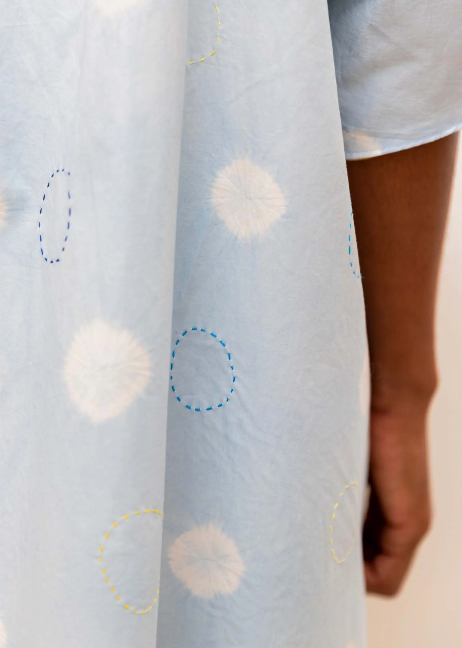 Close-up of a person wearing a light-colored shirt with circular patterns in white, yellow, and blue, standing near a wall and a door.