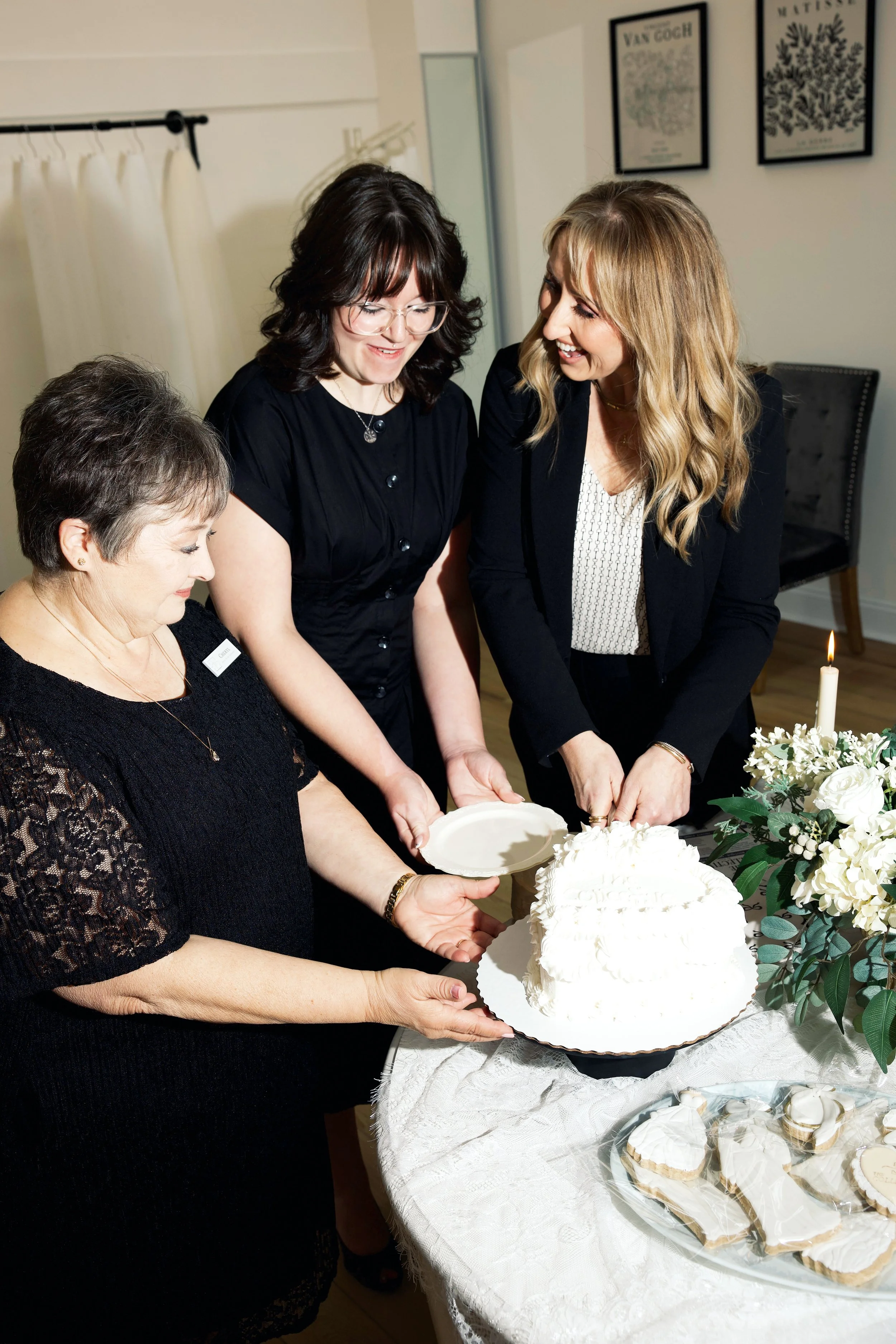 Three women are cutting a white cake at a celebration, with a table of cookies, cupcakes, and floral arrangements nearby.