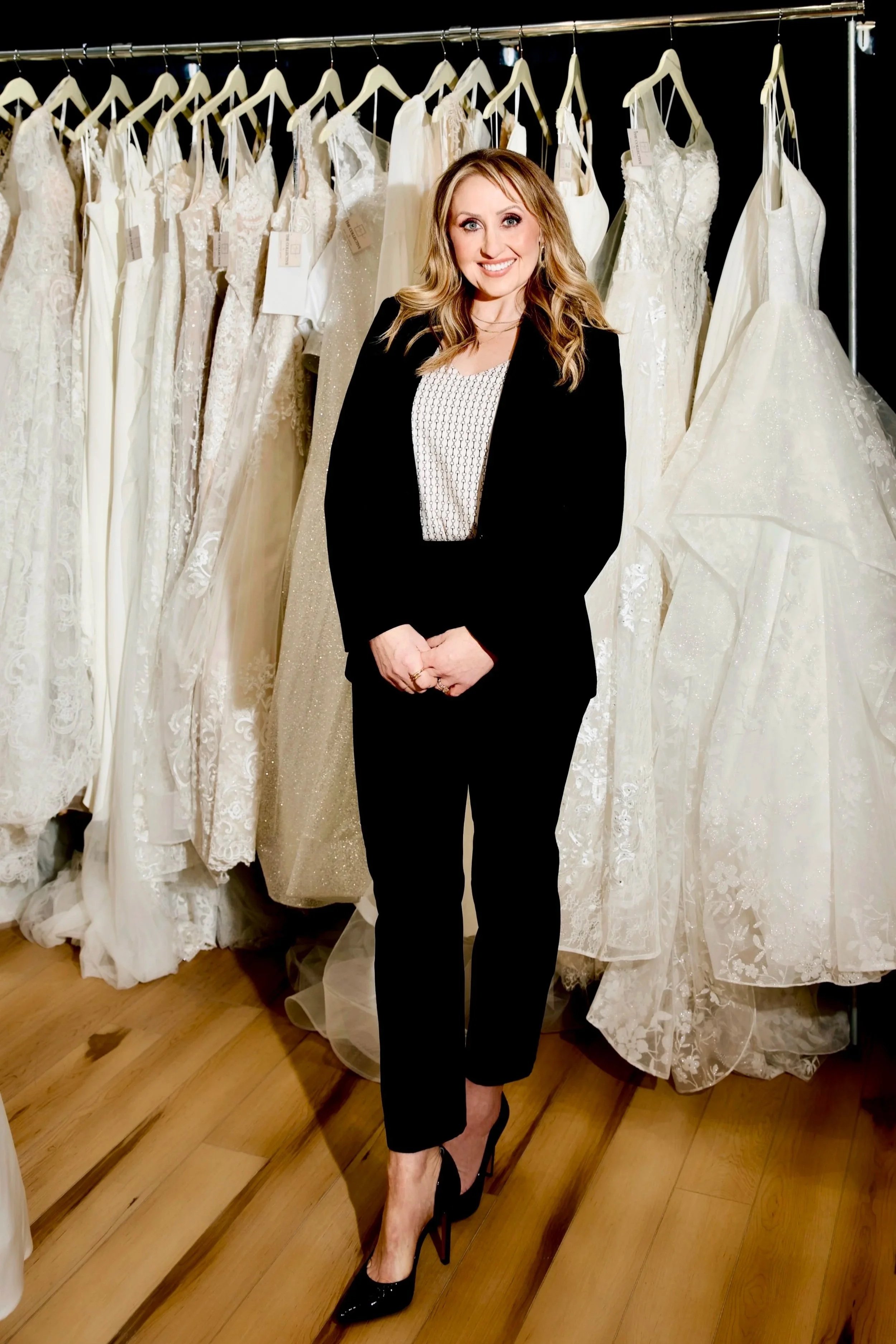 A woman in black pants and a black blazer stands in front of a rack of white wedding dresses, smiling at the camera.