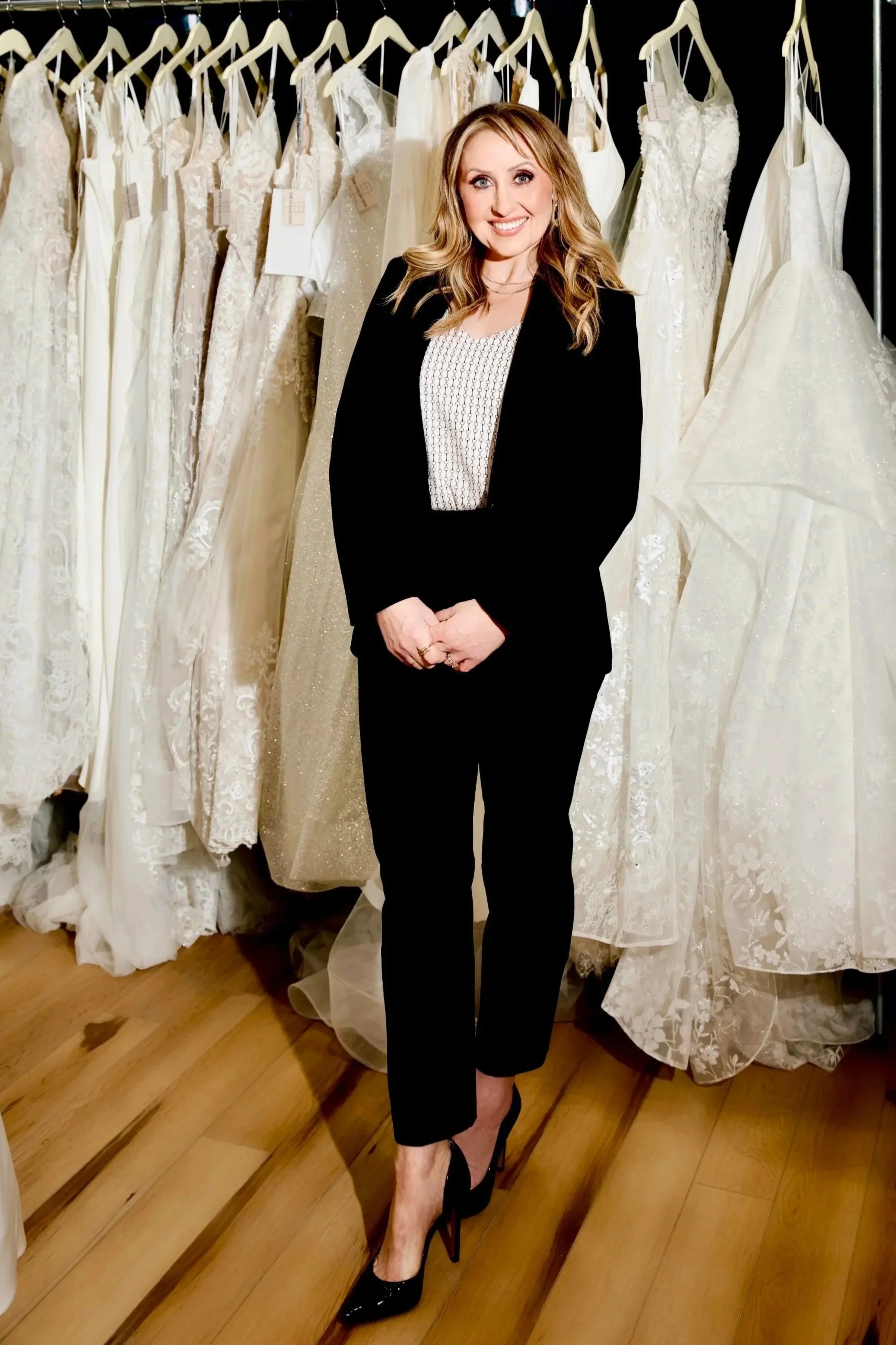 A smiling woman dressed in black pants and a black blazer over a white patterned top, standing in front of a rack of wedding dresses in a bridal store.