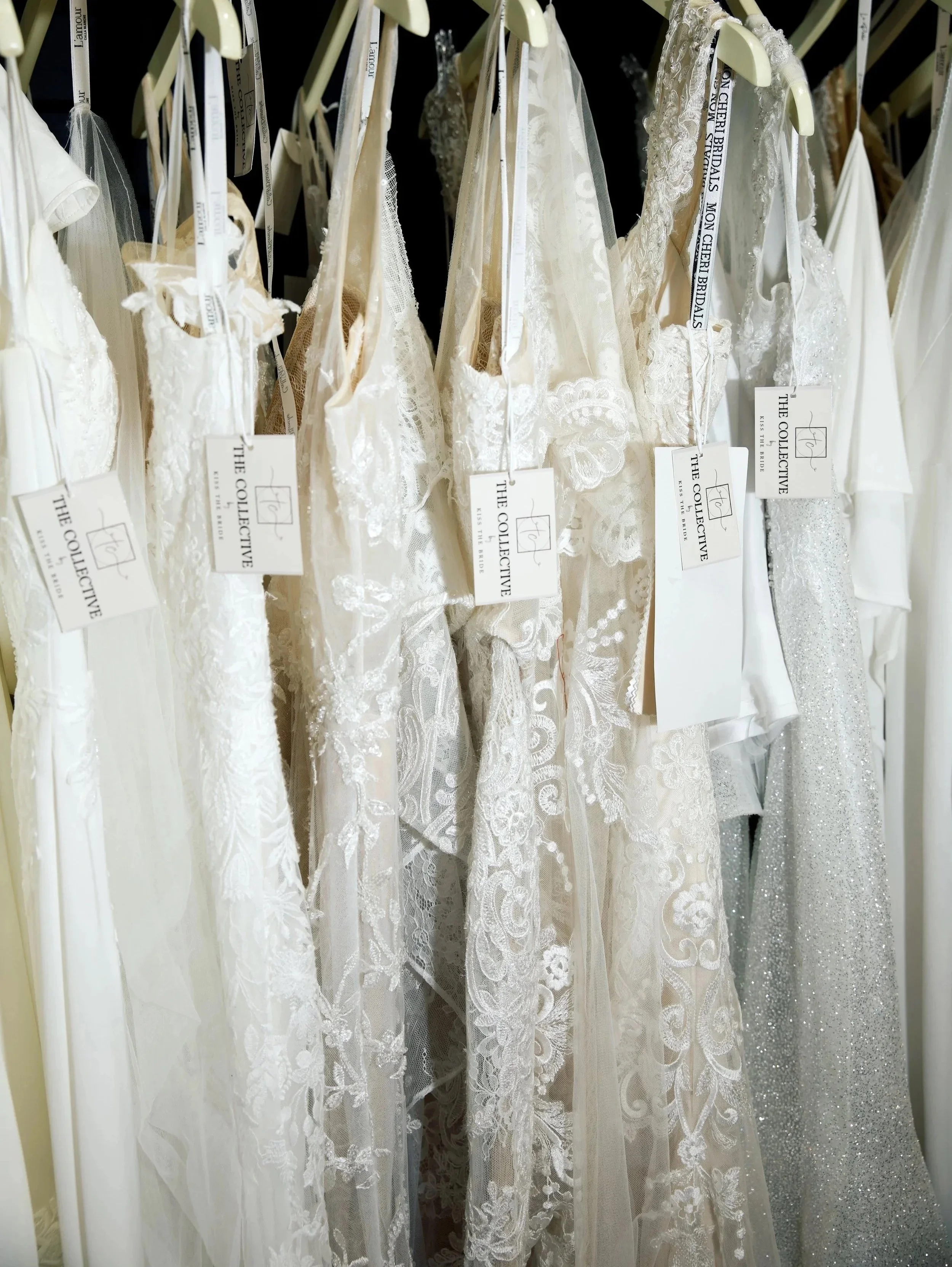 A row of white bridal dresses hanging on beige hangers in a store. The dresses feature various lace and embroidery designs, some with tulle and beaded details. The tags attached to the dresses read 'The Collective' and 'KISS THE BRIDE'.