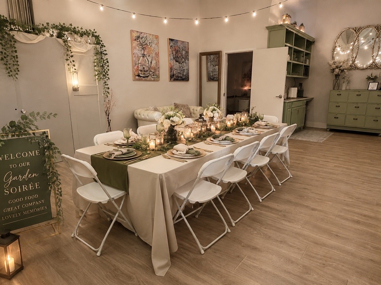Large dining table set with plates, napkins, glasses, and a green and cream table runner, decorated with candles and floral arrangements, in a warmly lit room with string lights, framed artwork, and green furniture.