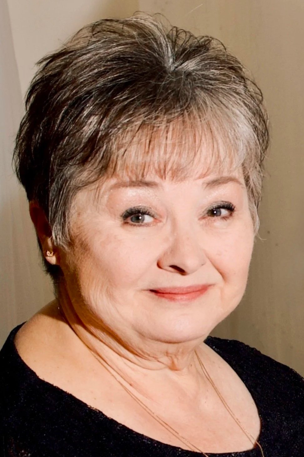 Close-up portrait of an elderly woman with short, layered gray hair, wearing a black top and a delicate gold necklace.