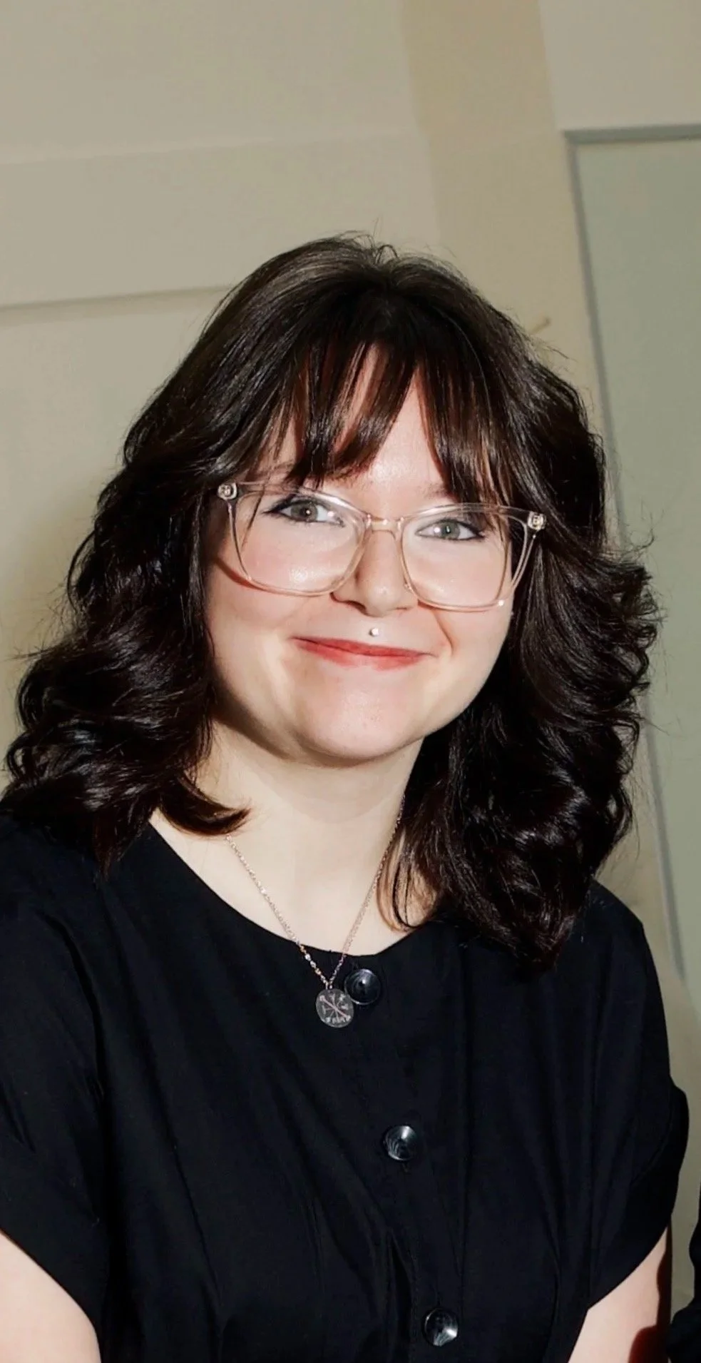 A woman with shoulder-length dark curly hair, wearing glasses, a black top with buttons, a necklace, and a facial piercing on her upper lip, smiling at the camera.