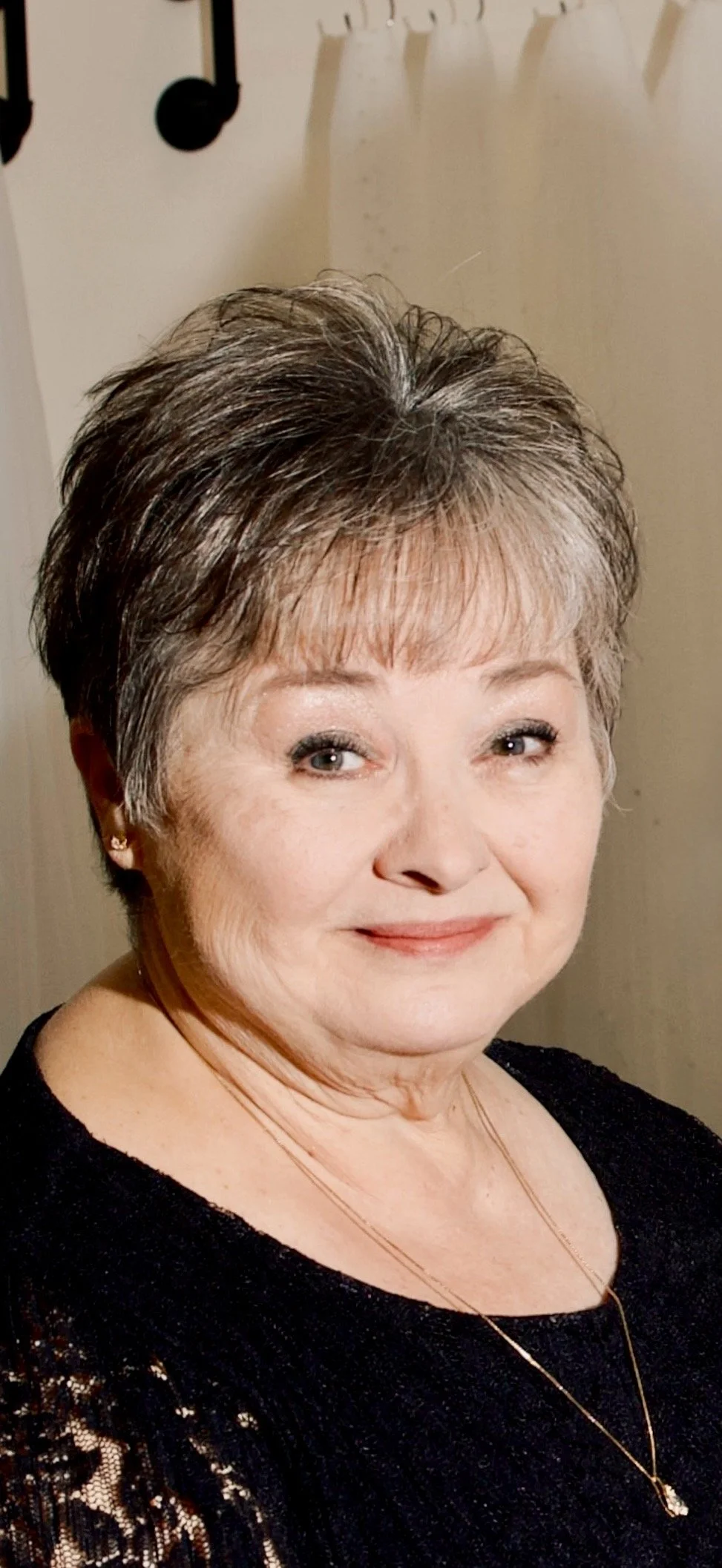 A close-up portrait of an older woman with short, dark, and gray hair, smiling, wearing earrings and a black lace top, standing in front of a light-colored wall with hooks.