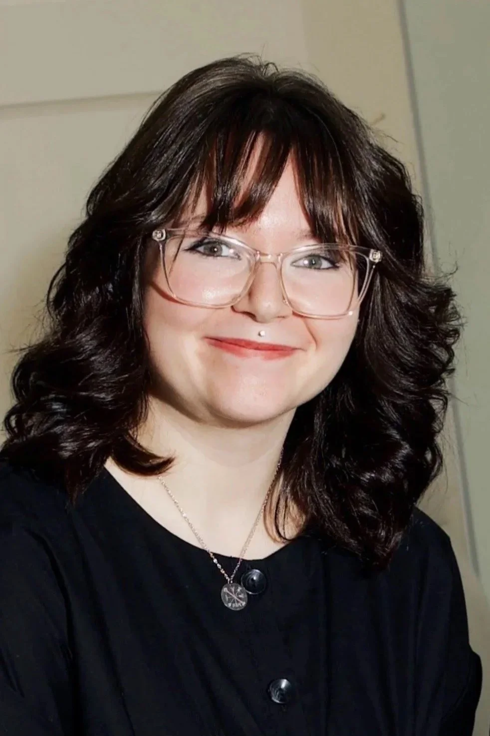 A woman with dark, curly hair and glasses, smiling, wearing a black top and a necklace with two pendants.