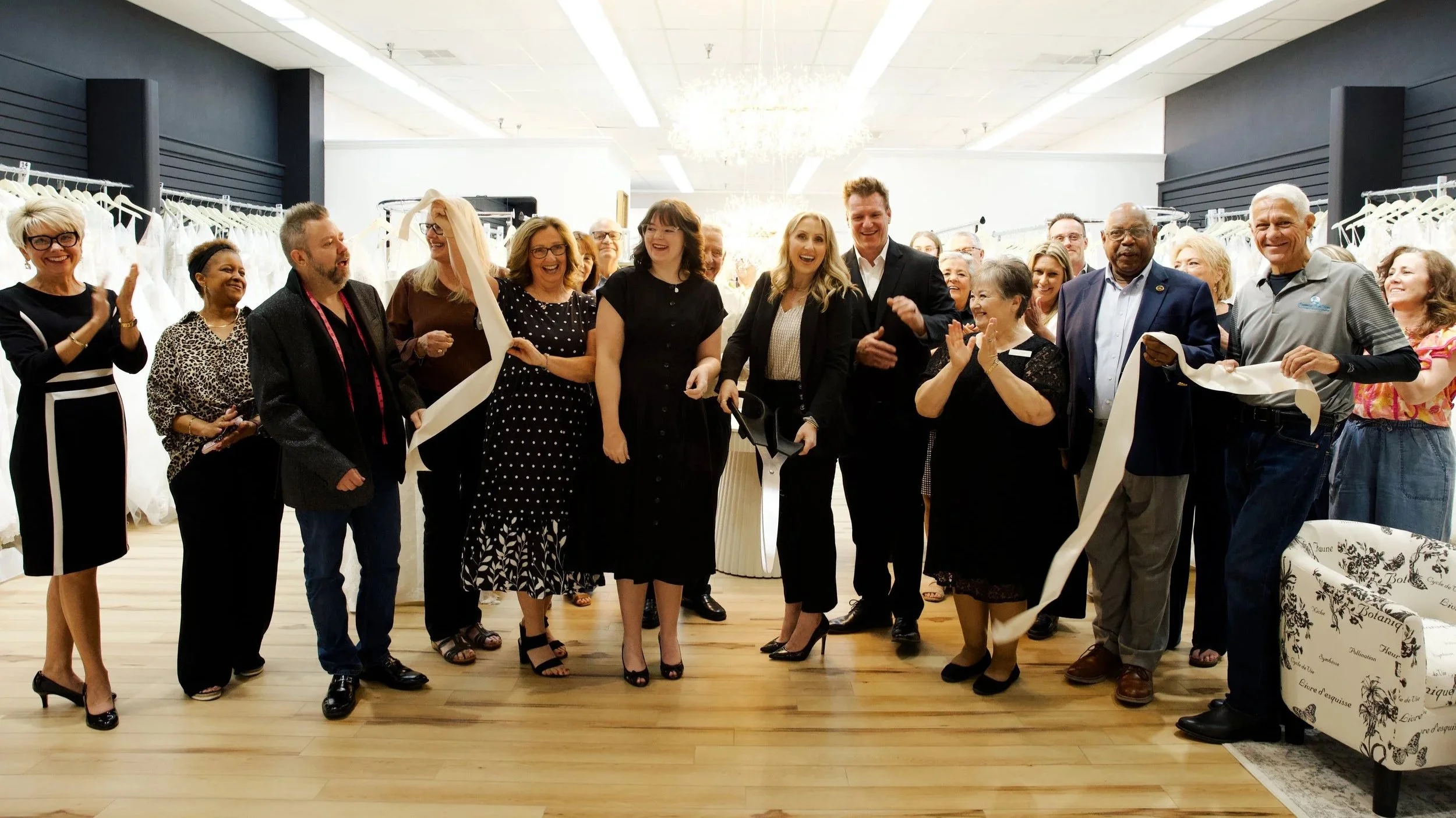 Group of people at a ribbon-cutting ceremony, some clapping and smiling, in an indoor event space with racks of clothing in the background.