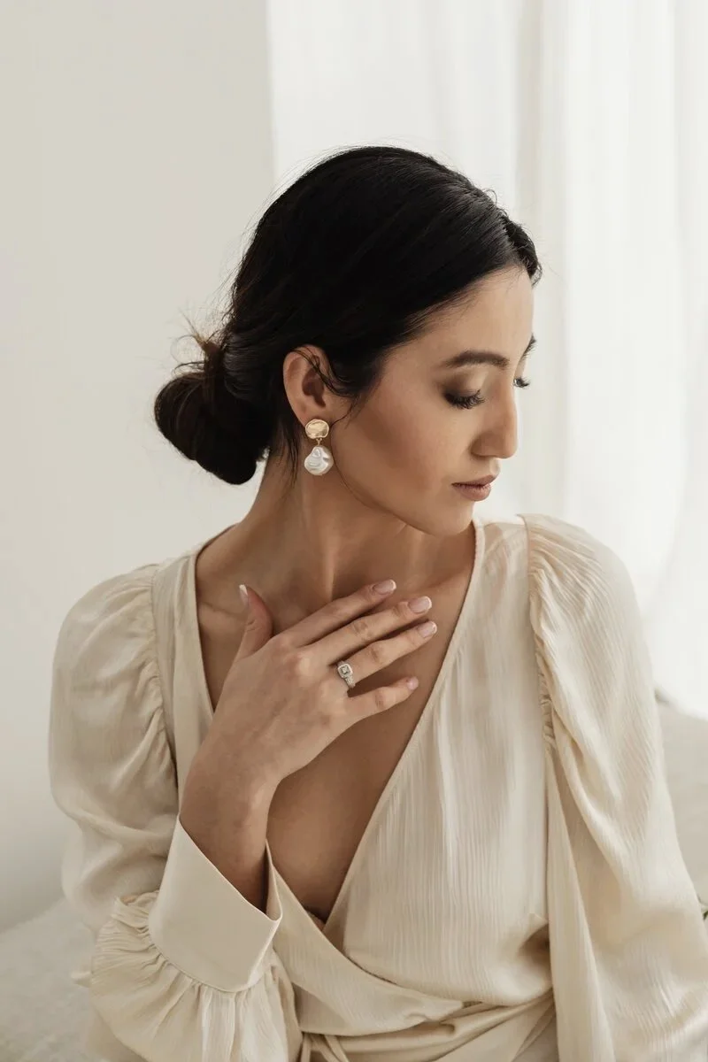 A woman with dark hair styled in a low bun, wearing pearl and gold earrings, and a ring on her finger, is gently resting her hand on her chest while looking down, dressed in a cream-colored, textured blouse with puffed sleeves, in front of a light background.