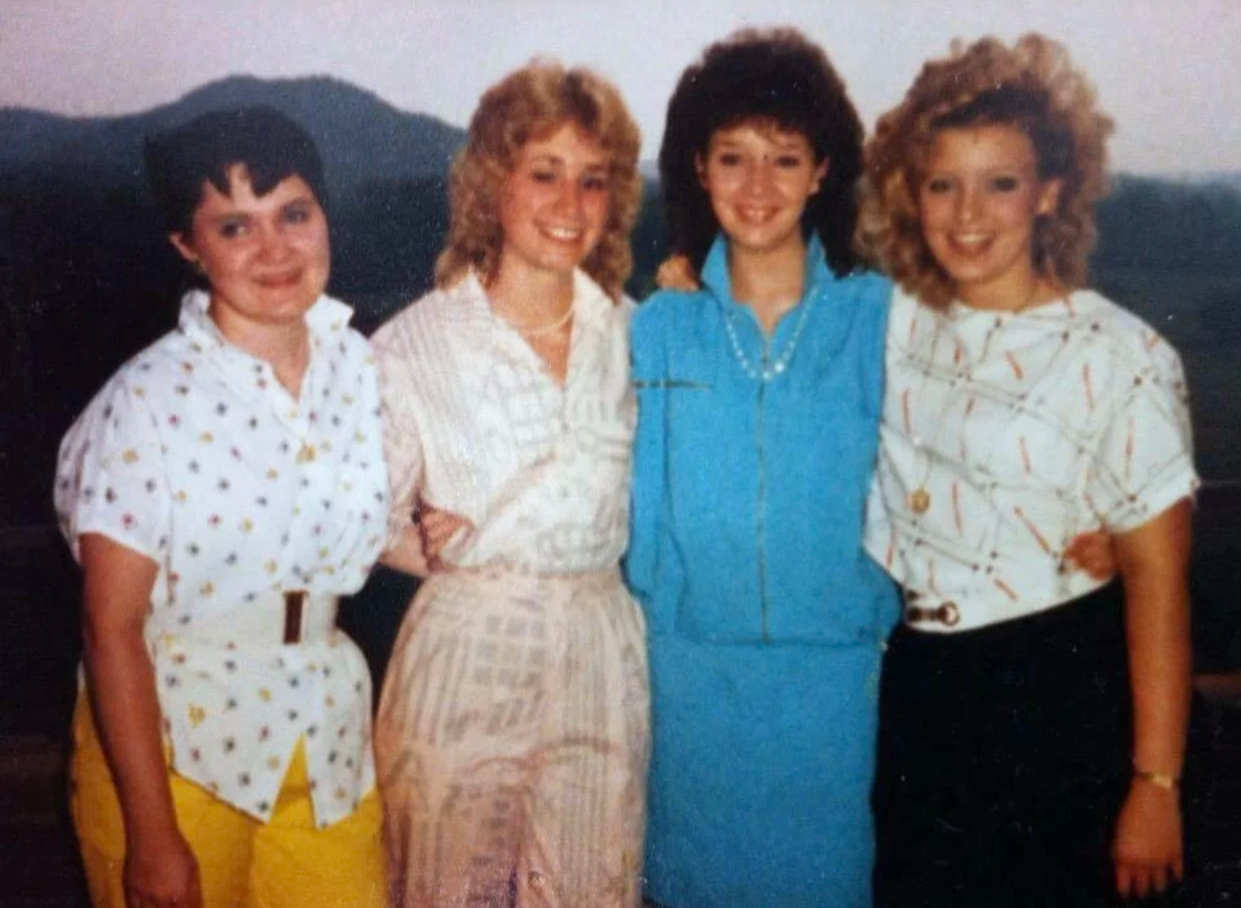 Four women standing outdoors with mountains in the background, wearing 1980s casual clothing and smiling.